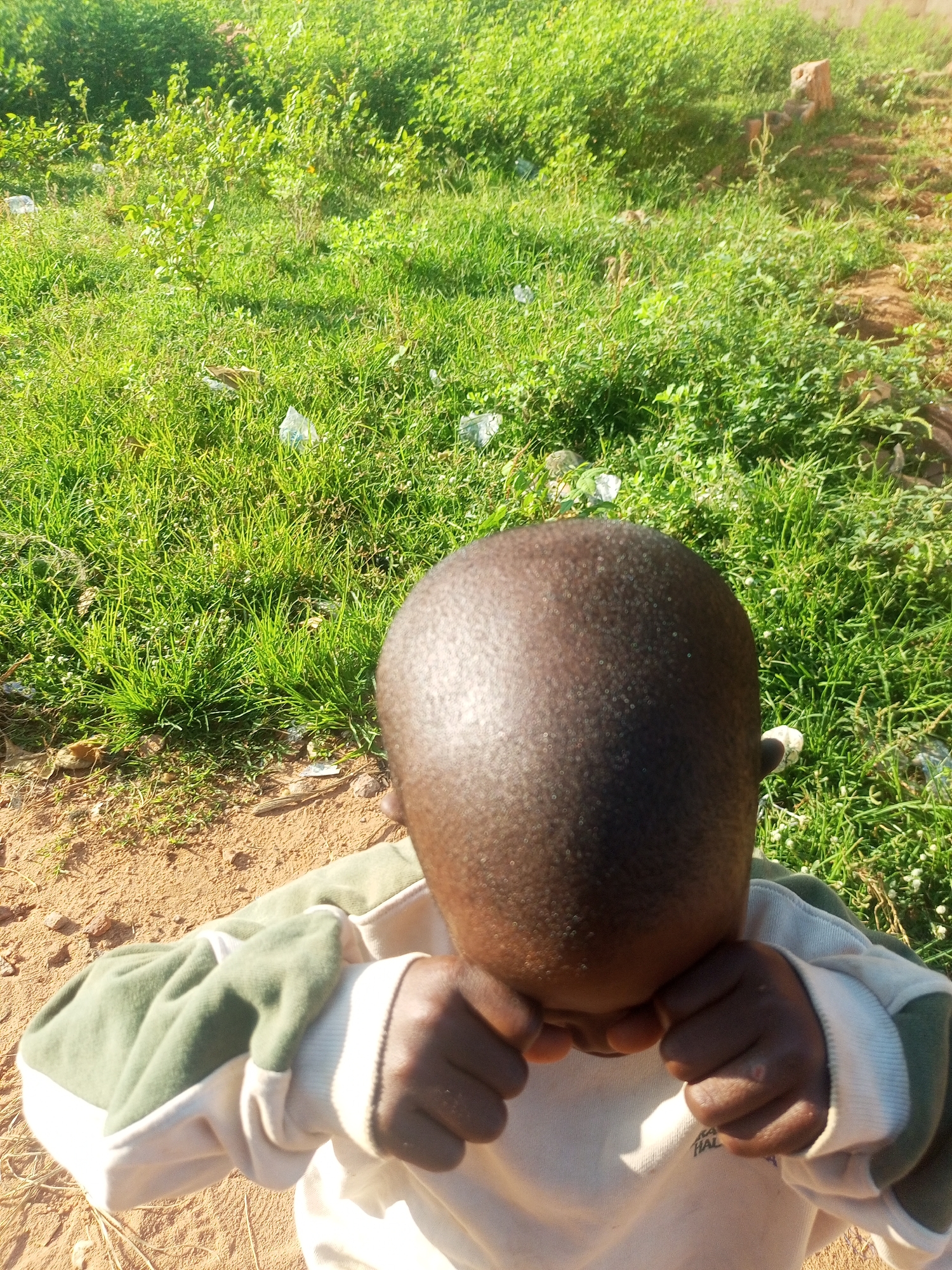 A child is facing away from the camera, with their head bowed and their face obscured by their arms. They are wearing a light-colored sweatshirt with green sleeves. The child is standing on a dirt path, with a grassy area behind them. The environment appears to be an outdoor setting, possibly a park or a yard, with sunlight casting shadows. There are some small white objects scattered in the grass, which could be litter. The background shows more green vegetation and some reddish-brown soil. The image does not provide any specific clues to pinpoint the location within Tempure, Nigeria.
