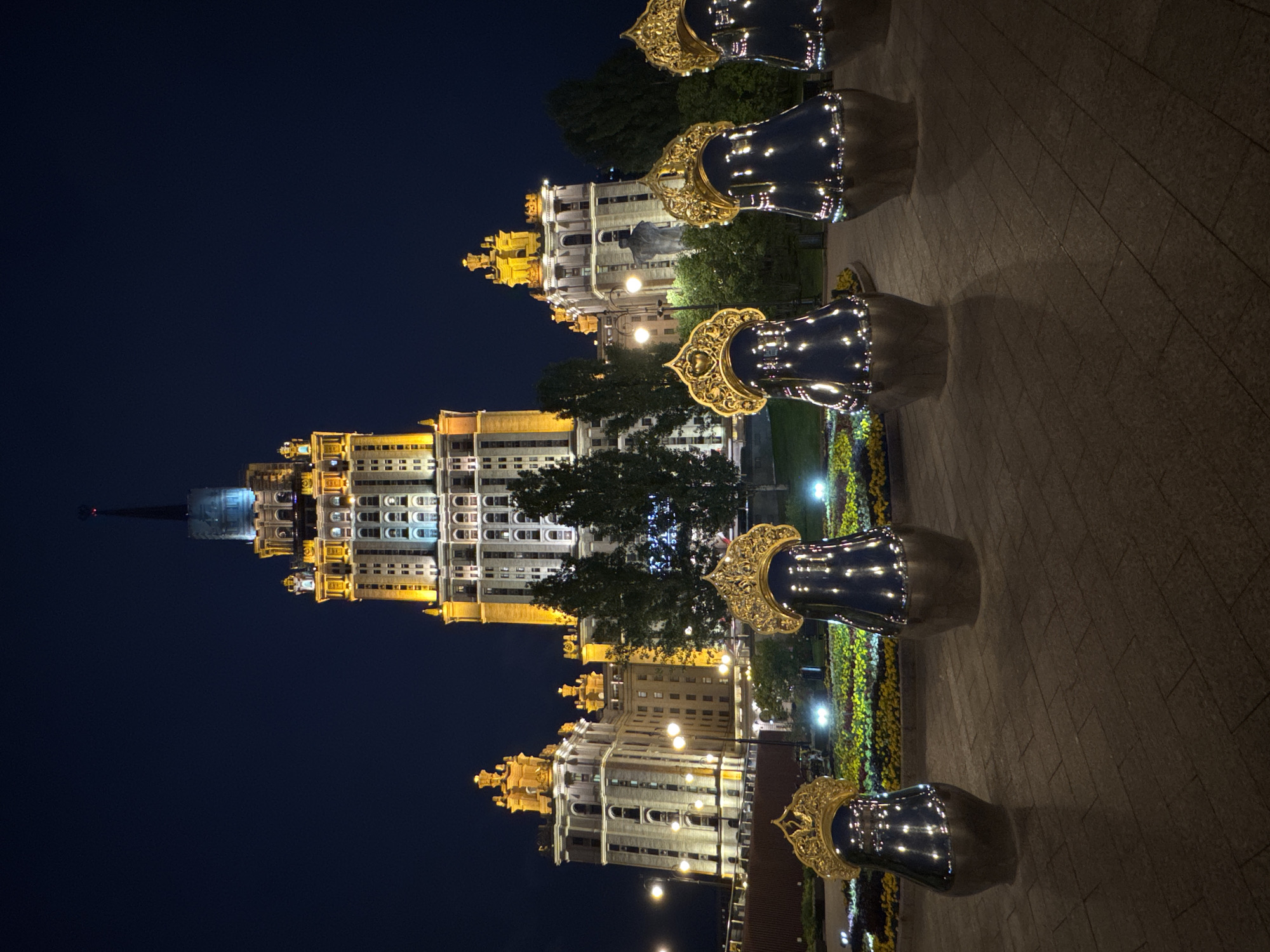 A night-time, low-angle exterior, wide shot captures the illuminated Stalinist skyscraper, the main building of Moscow State University, and its surrounding architectural elements, as well as a row of large, reflective, matryoshka doll-shaped sculptures in the foreground. The skyscraper, with its prominent spire, is lit with warm yellow and white lights, highlighting its many windows and decorative features. To the left and right of the central building are other illuminated structures with similar architectural styles, featuring golden accents. The sky is a deep, dark blue, indicating it is nighttime. In the middle ground, trees are visible, and below them, a flowerbed with yellow and pink flowers is softly lit. The foreground features a paved walkway made of rectangular stones, upon which stand six large, dark, reflective sculptures shaped like matryoshka dolls, each topped with a golden, ornate crown-like headdress. The sculptures reflect the surrounding lights, creating a glittering effect. No people are visible in the image.