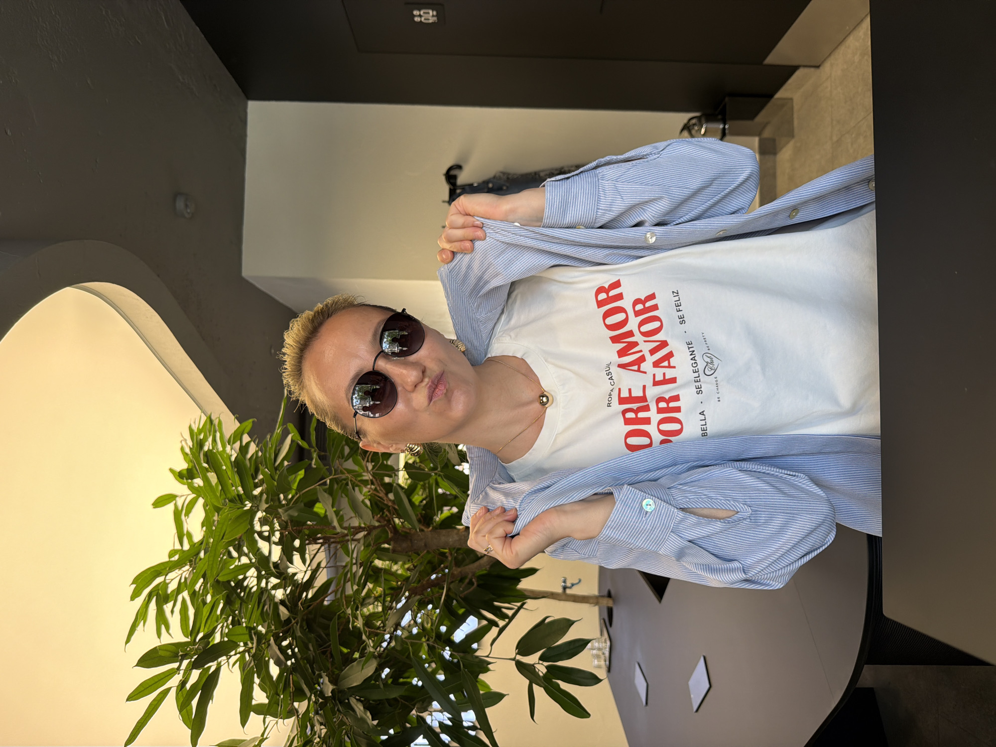 A woman with short blonde hair and sunglasses stands in a room with a large green plant. She is wearing a white t-shirt with the words "ORE AMOR POR FAVOR" in red letters and a blue and white striped shirt over it. She is holding the shirt open with her hands. The room has a gray and white wall and a dark gray table. The location of this photo cannot be identified.