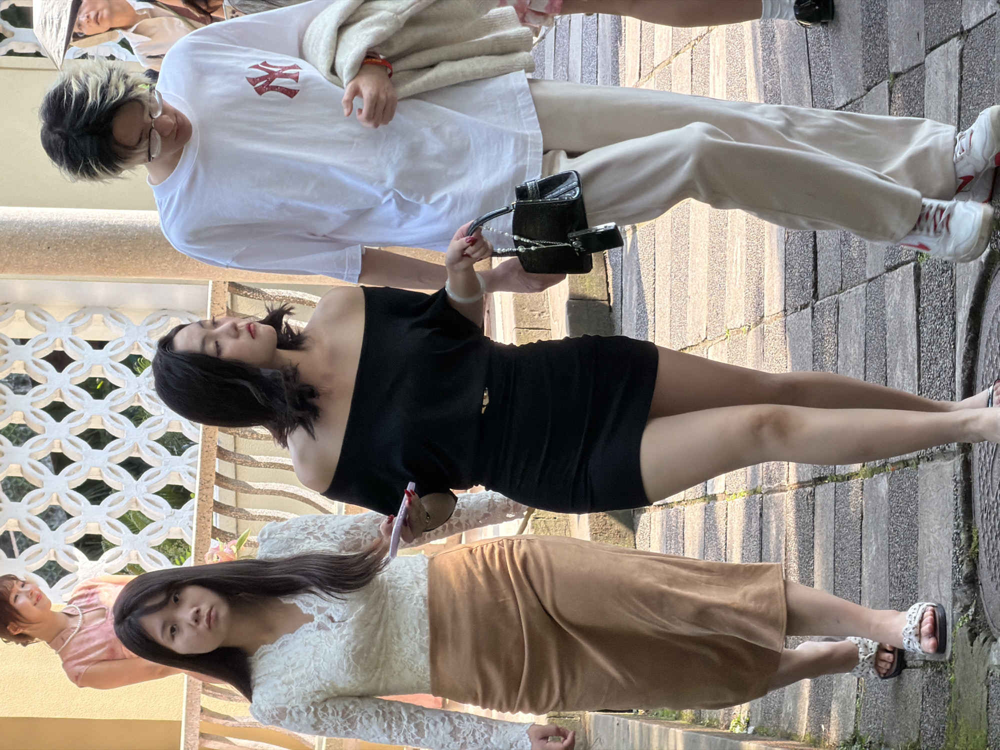 Three individuals are walking on a stone pathway in Jiangbeicheng Street, China. In the foreground, a woman on the left wears a white lace top and a tan skirt. She is walking towards the viewer. Next to her, a woman in a black off-the-shoulder dress walks in the same direction, carrying a black handbag with pearl embellishments. To the right, a person with black and blonde dyed hair wears a white t-shirt with a red logo and beige pants. They are looking down and walking away from the viewer. In the background, another woman in a pink dress and a pearl necklace is visible. The pathway is bordered by a decorative stone railing and foliage. The lighting suggests it is daytime.
