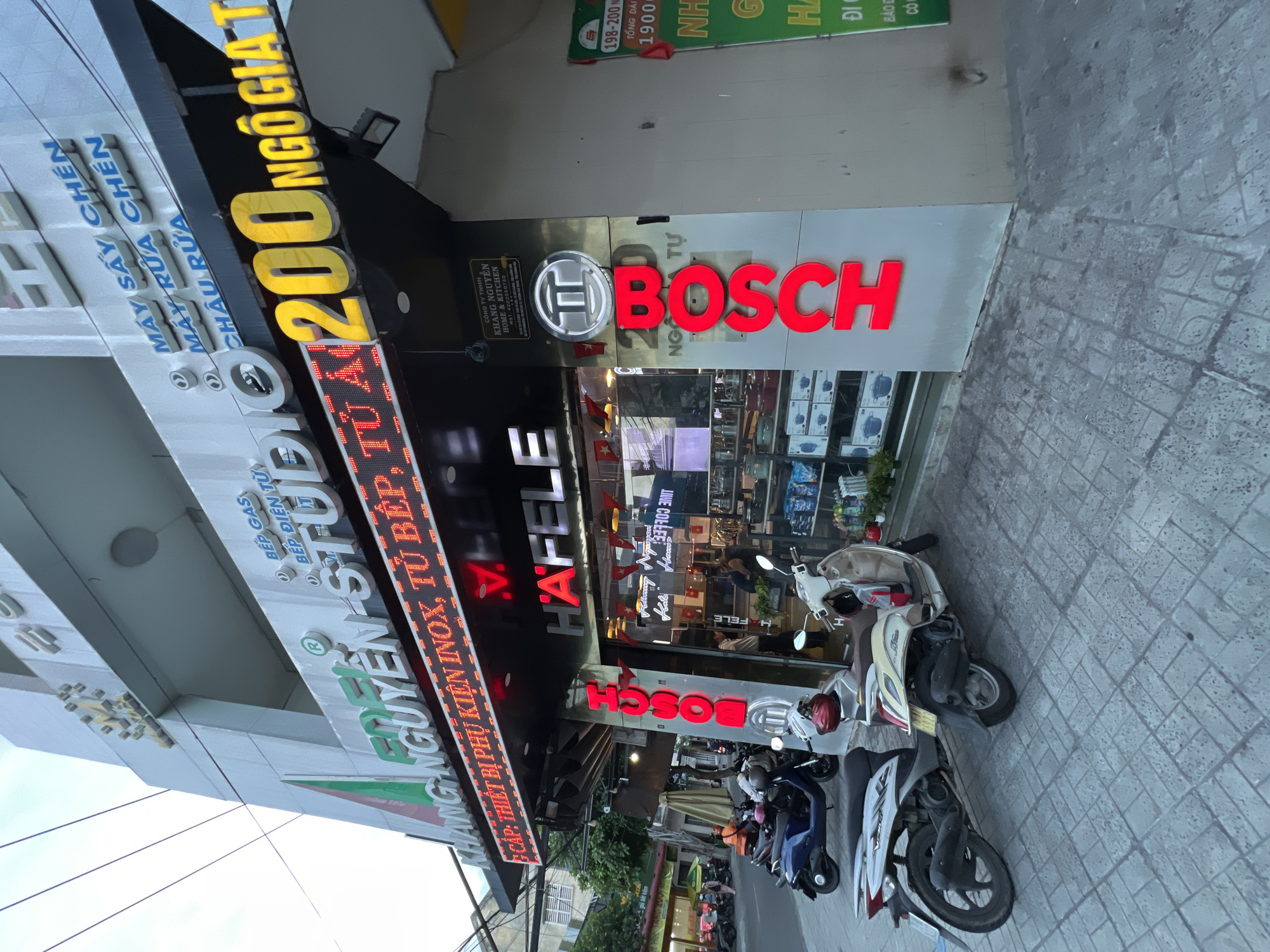 The image captures a street view of a store called "KHANG NGUYEN STUDIO" located at "200 NGO GIA TU" in Nha Trang, Vietnam. The storefront features prominent signage in Vietnamese and English, advertising kitchen appliances and accessories. A large red "BOSCH" logo is visible on the side of the building, along with "HAFELE" signage. Several motorcycles are parked on the sidewalk and street in front of the store. The building appears to be a multi-story structure, typical of urban commercial areas in Vietnam. The sky is overcast, suggesting it might be a cloudy day.