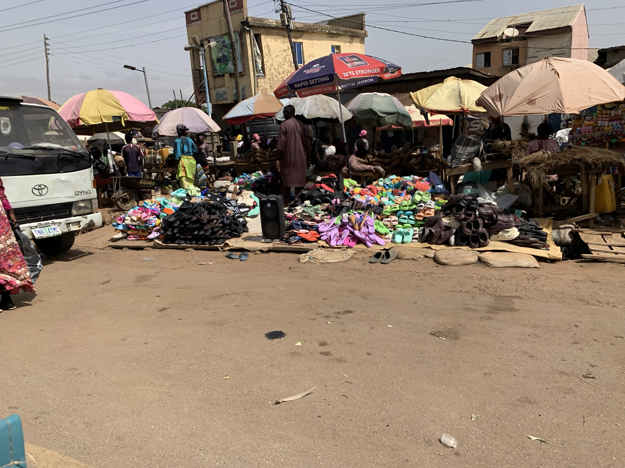 The image depicts a bustling market scene in Rafin Par, Nigeria. A large assortment of colorful sandals and shoes are piled in the foreground, partially obscuring the vendors seated behind them under a variety of colorful umbrellas. People are milling about, presumably shopping. To the left, a white Toyota Dyna truck is parked, its license plate visible. The background reveals a mix of buildings, some with signs, under a slightly overcast sky. The road in the foreground shows signs of wear and scattered debris.