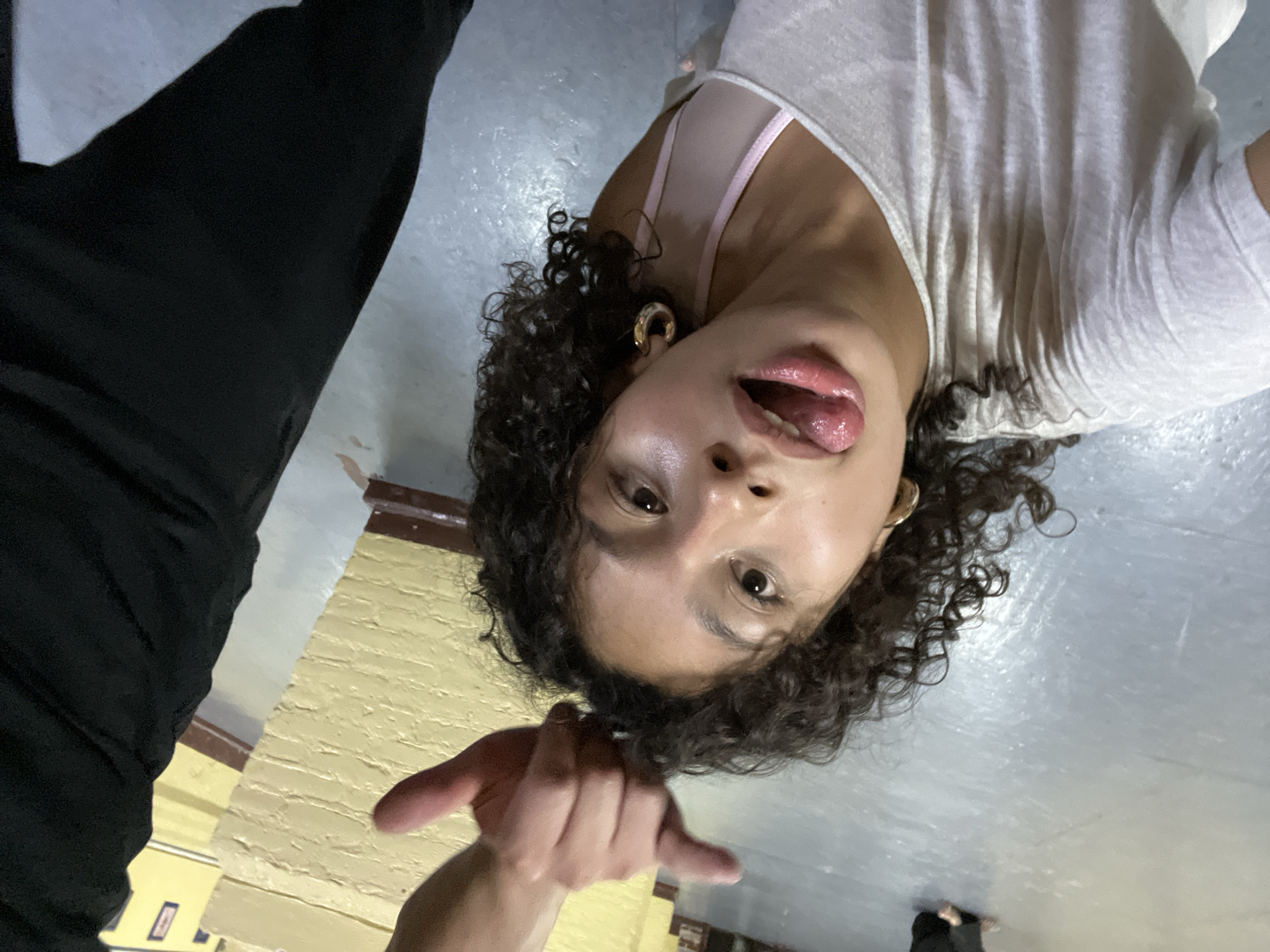 A photograph, shot from a low angle, depicts a person with curly dark hair, fair skin, and brown eyes lying on their back on a gray tiled floor. The person is wearing a light-colored v-neck shirt and a pink top underneath. The person's tongue is sticking out, and their mouth is open. A hand with a pointing finger is visible in the foreground. The background features a wall painted in a yellow hue. The location is Unknown, Unknown.