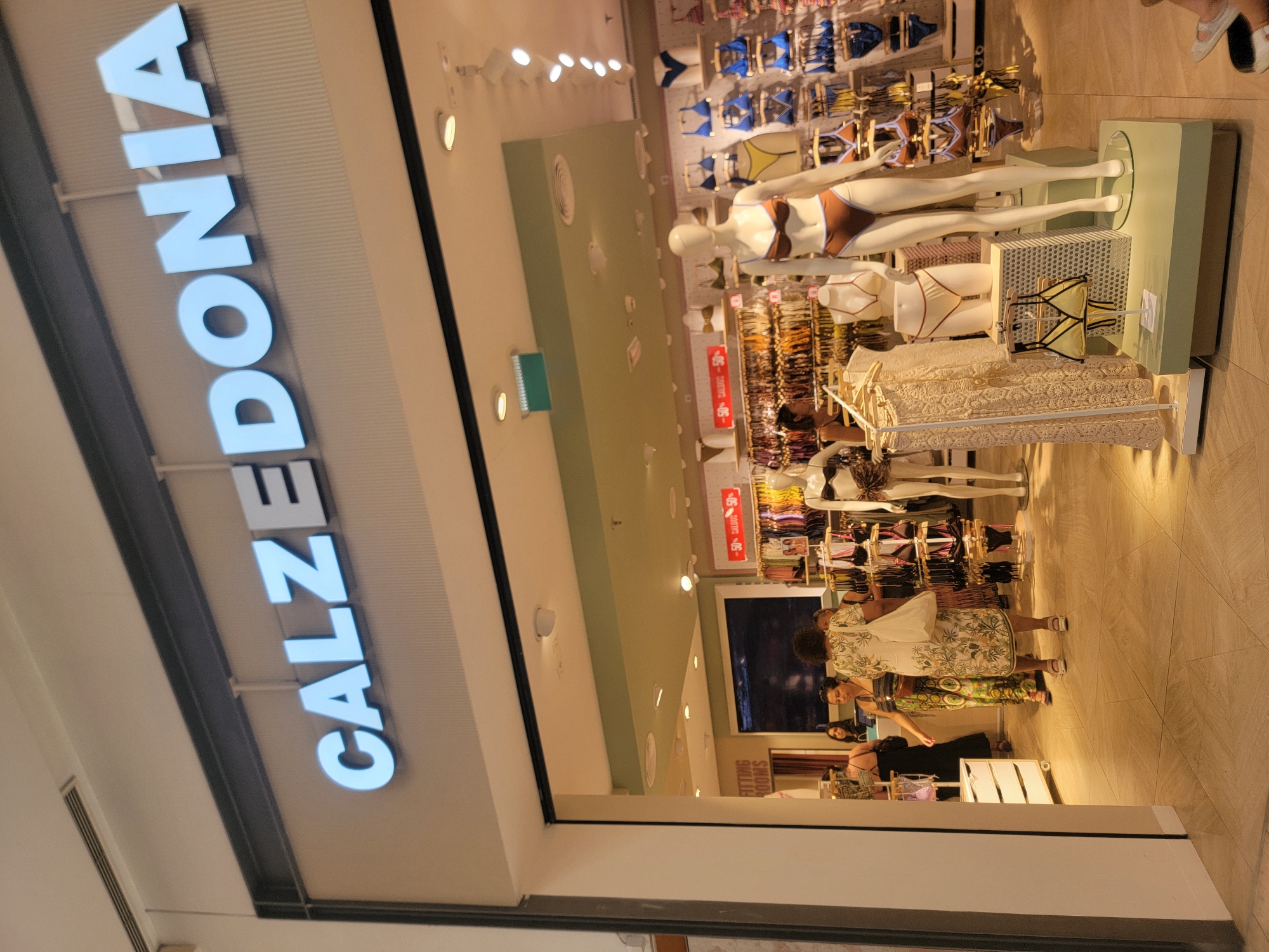 The image shows the exterior of a Calzedonia store, a retail establishment. The store's name, "CALZEDONIA," is prominently displayed in large, illuminated blue letters above the entrance. Inside the store, visible through the open doorway, racks of clothing, mannequins displaying swimwear, and a sales counter are discernible. A few customers are browsing or interacting within the store. The flooring appears to be tiled. The environment suggests an indoor shopping mall setting. The image has been identified as being taken in Faro, Portugal.