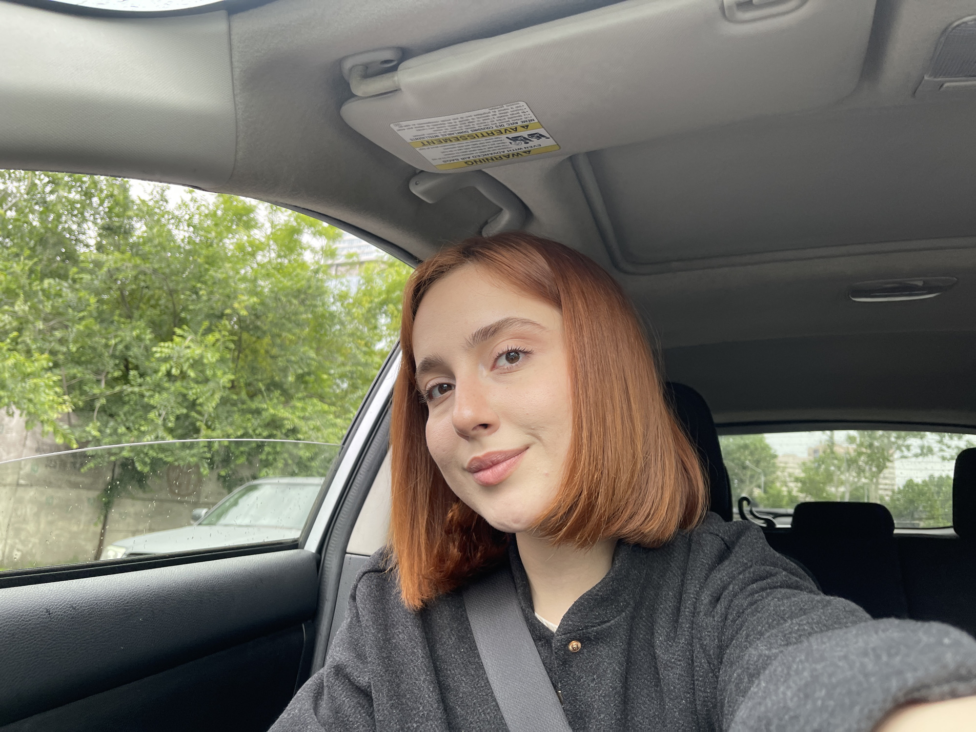 A young woman with red hair is in the driver's seat of a car, wearing a seatbelt and a gray jacket. The car's interior is visible, including the dashboard, sun visor, and seats. Through the side window, trees and a building are visible, suggesting an urban environment. Based on the architecture and vegetation, the location is plausibly Tbilisi, Georgia. The image appears to be a selfie taken by the woman.