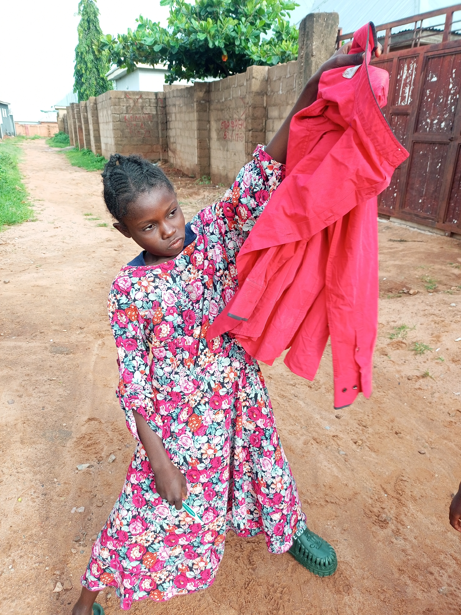 A young girl stands in a sandy, unpaved alleyway in Tempure, Nigeria, holding up a bright red shirt. She is wearing a floral-patterned dress with pink, white, and green flowers. Her hair is braided and pulled back. The shirt she is holding is a button-down style with the collar visible. In the background, there is a brick wall and a red metal gate. The ground is sandy with patches of dry grass.