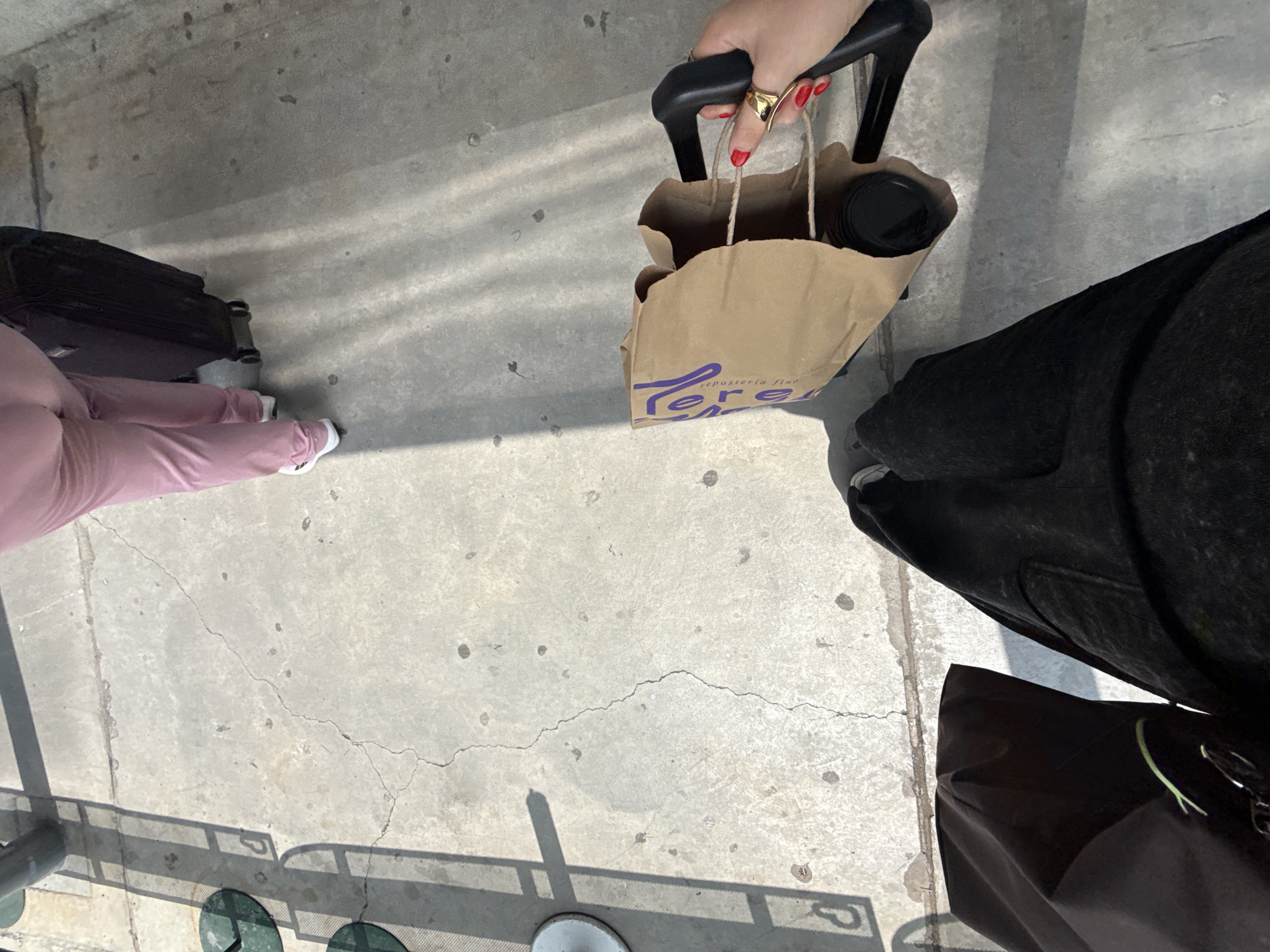 The scene appears to be on a concrete outdoor surface in Apodaca, Mexico. A person wearing pink pants stands next to a black suitcase with wheels. Another person is partially visible wearing black clothing and black shoes, carrying a brown paper bag with the logo "Tere Reposteria Fina" visible. The person holding the bag has red nail polish and a gold ring on one hand, holding the retractable handle of the bag. Another dark-colored bag is also on the ground beside the person in black. A metal railing with circular green and white elements is visible at the bottom of the scene.