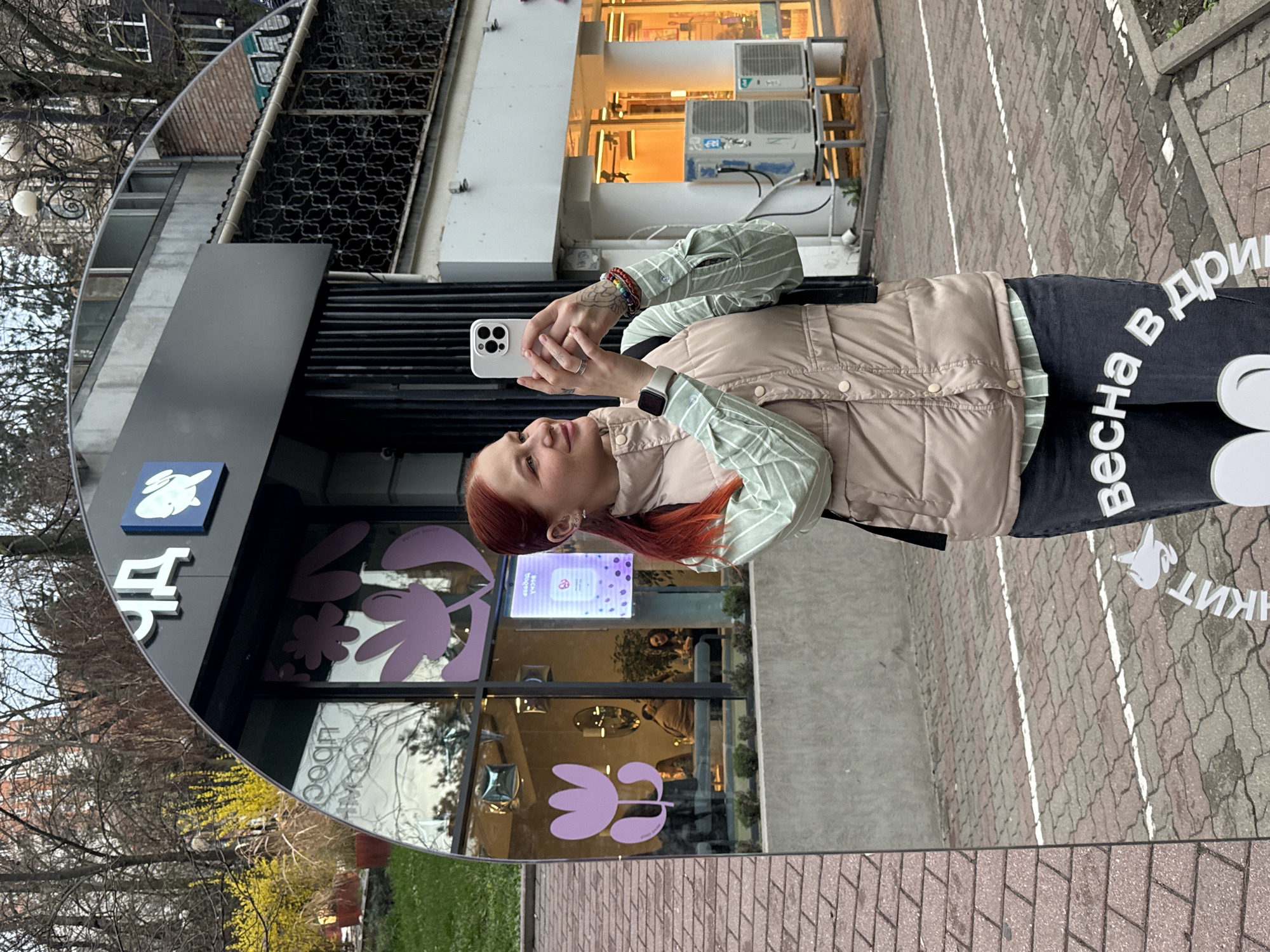 A woman with red hair is reflected in a round mirror outside a building. The mirror reflects a florist shop. The woman holds a white smartphone. She wears a light tan puffer vest, a green striped shirt, and a watch on her left wrist. The reflection shows store signage with purple flower motifs. Part of the sign says "Весна в дри". The surrounding environment shows a brick-paved street, building facades, and spring foliage. The location is likely in Ovoshchnoy, Russia, based on the Cyrillic lettering.
