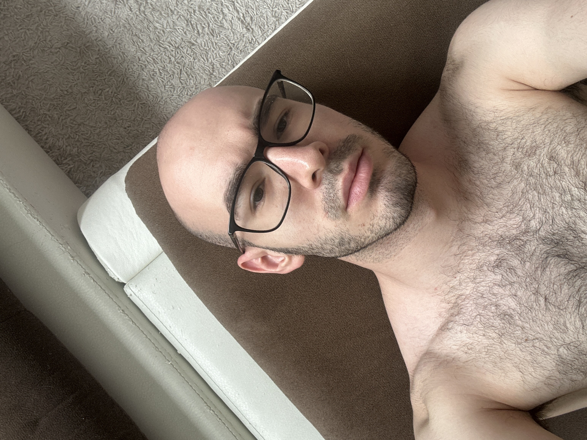A close-up, top-down shot depicts a fair-skinned, bald man with stubble and black-framed glasses. He is lying on a brown and white couch. The man's chest, covered in dark hair, is visible. The background includes a portion of a gray carpet and the off-white material of the couch. There is no indication of the location as Moscow, Russia or any other city.