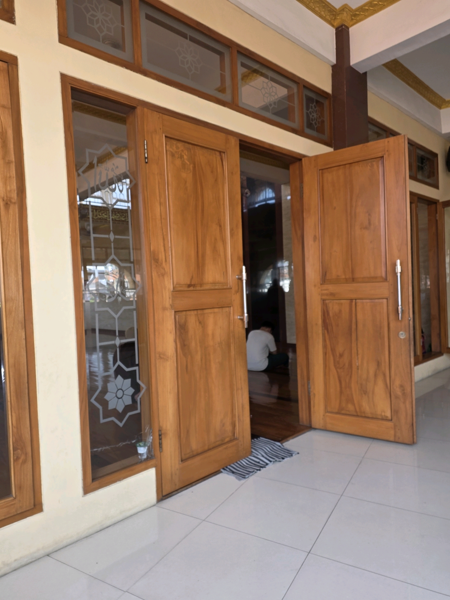 A side view shows an open wooden door revealing a person sitting in prayer within a room. The interior of the room has a polished wooden floor and patterned glass windows. The exterior features a light-colored wall and a tiled floor. The overall scene suggests an indoor setting, possibly a home or a place of worship, located in Bojongsoang, Indonesia.