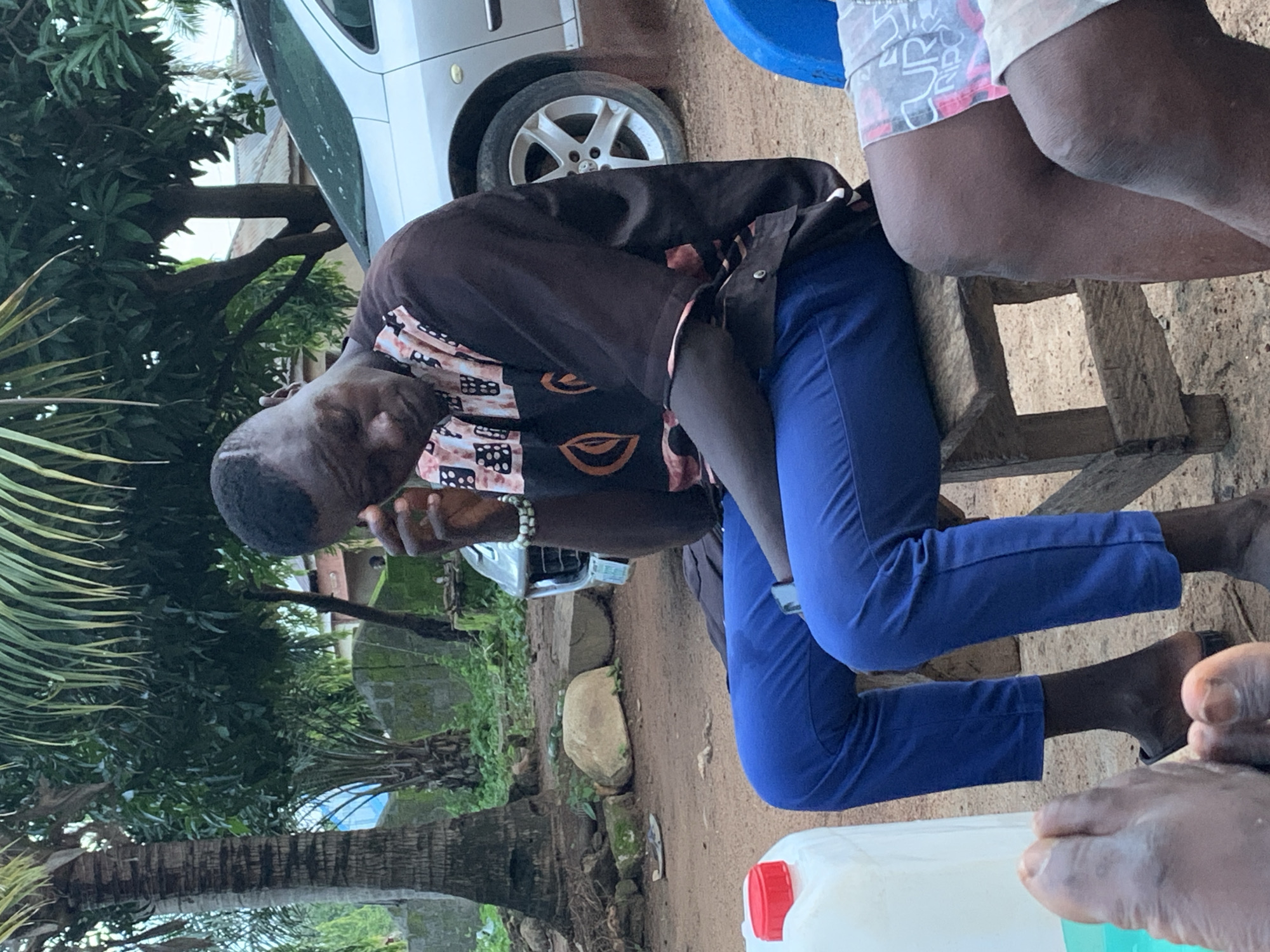 A man with dark skin and short hair sits on a wooden stool outdoors, with his head bowed and his right hand touching his forehead. He wears a dark brown shirt with a patterned design in black, white, and orange on the sleeves and chest, and bright blue trousers. A silver car is visible behind him. In the foreground, a large white plastic container with a red lid is partially visible on the left. The ground is dirt and gravel, with some green foliage and trees in the background. The setting appears to be a residential area in Akwanga, Nigeria.