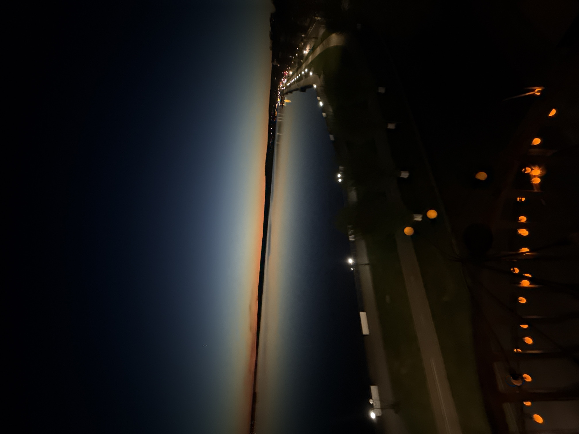 At dusk in Votkinsk, Russia, the sky transitions from deep blue to orange near the horizon. A body of water reflects the fading light. Along the shore, evenly spaced streetlights illuminate a pathway. Further in the distance, more lights suggest a town. The foreground is dark, with a vertical string of orange lights. The lights appear to be hanging from a structure, possibly a building or fence.