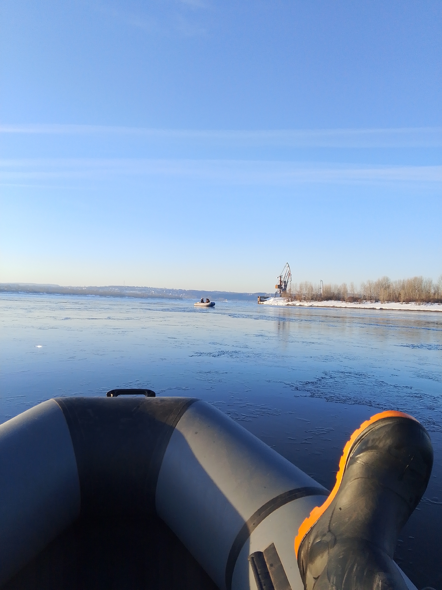 A wide-angle shot shows a partial view of a gray inflatable boat, with a black boot and orange sole resting on the side, in a partially frozen body of water. In the background, a crane is visible on the shore, along with bare trees, suggesting a cold climate. Another small boat with people is visible further out on the water. The sky is blue with light clouds. The scene is located near Chaikovsky, Russia, based on the visible landscape and buildings.