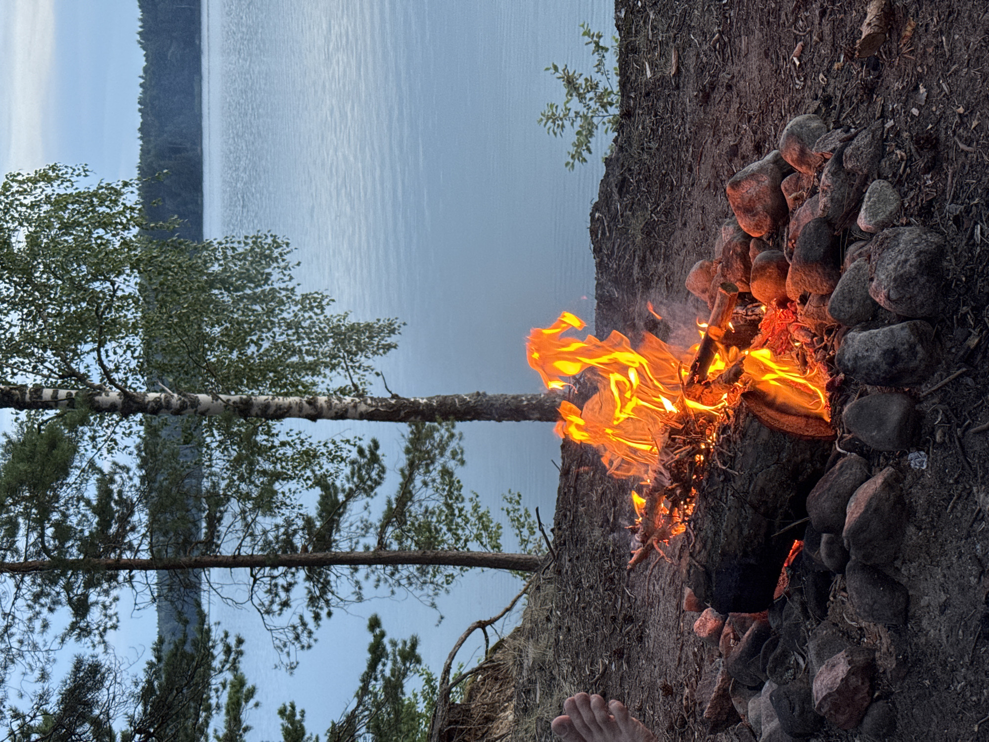 A campfire burns brightly on a rocky shore overlooking a calm, blue lake. The scene is set in a natural environment with trees lining the bank, their branches reaching towards the sky. A human foot is visible in the lower left corner of the frame, close to the fire. The fire is contained within a ring of stones, with logs and branches fueling the flames. The water appears still, reflecting the soft light of the sky. The overall impression is one of a peaceful outdoor setting.