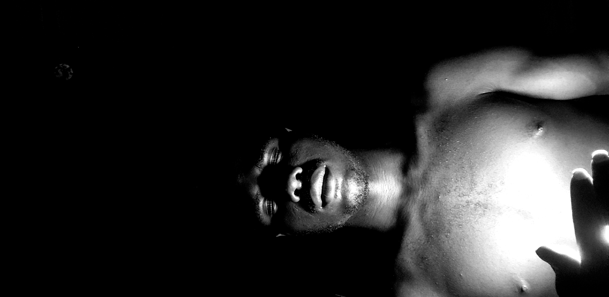 In a low-light, black and white image, a shirtless Black man's face and upper torso are visible against a dark background. The lighting creates strong shadows on his face, highlighting his eyes and mouth, which are slightly open. He appears to be looking upwards. His chest and shoulders are illuminated, revealing sweat or water droplets on his skin. His left hand is visible in the lower left corner of the frame, cupped as if holding a light source, which is the apparent source of illumination. The image is cropped in portrait orientation. There is no information within the image or the provided context that specifies a location in Narayi, Nigeria.