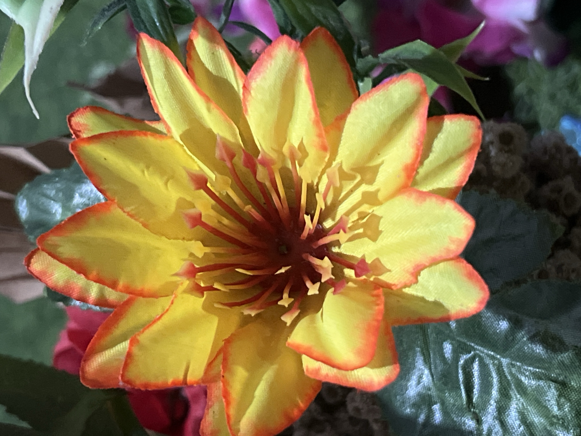 A close-up, high-angle, outdoor, natural light photograph captures a vibrant, artificial flower, likely part of a decorative arrangement. The flower is predominantly yellow with prominent red-orange petal edges and a deep red center surrounded by numerous delicate, thin, reddish-orange stamens. The petals are broad and layered, with visible texture suggesting they are made of fabric. Soft shadows are cast across the petals, indicating a light source from the upper left. The background is a blur of green foliage, hints of red and pink flowers, and some darker, possibly brown, elements, creating a sense of depth and a lush environment. The image was taken in Denpasar, Indonesia, and is a still life of decorative flora.