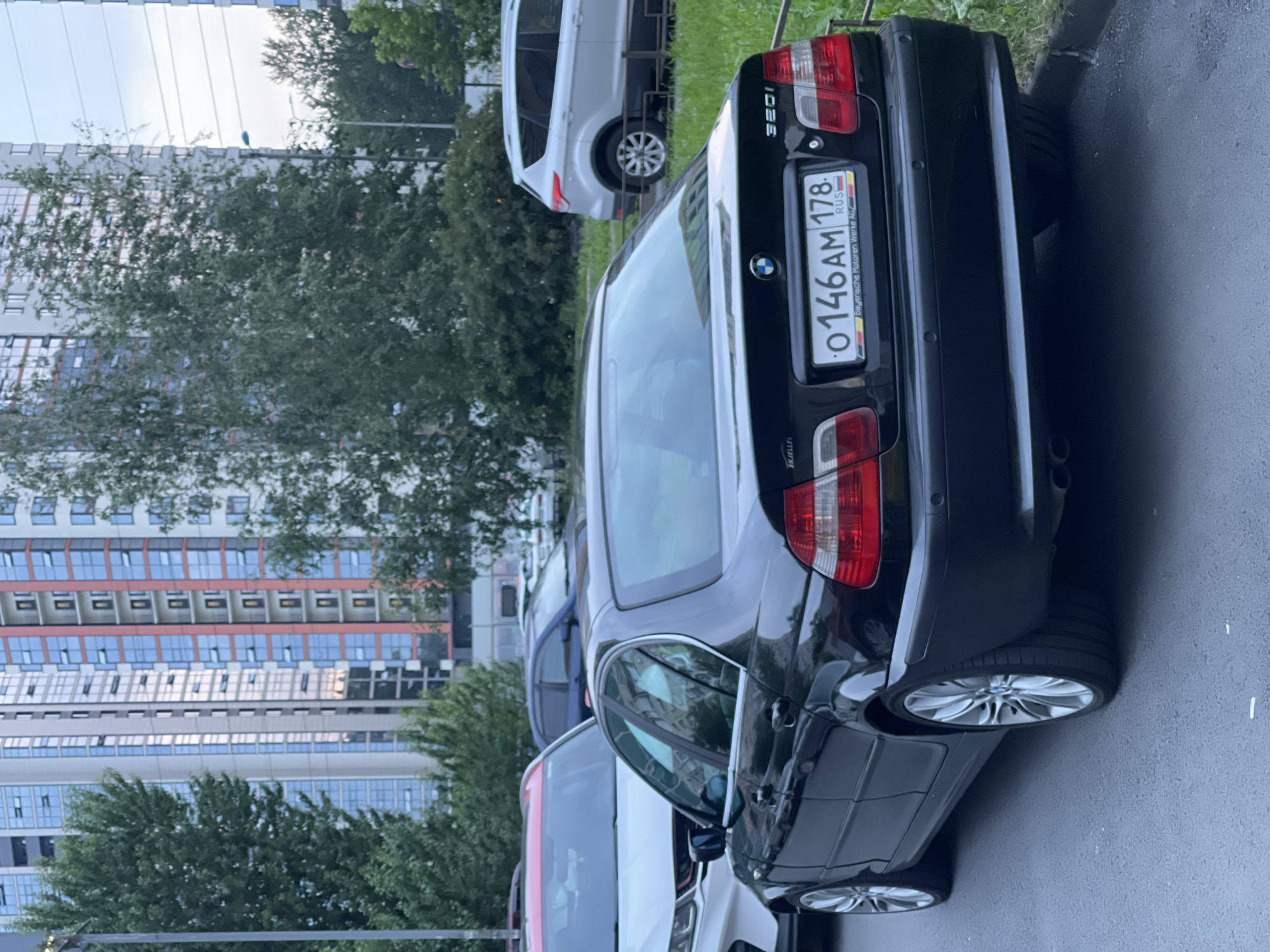 A black 2004 BMW 320i sedan is parked on a street next to a grassy area. The car's rear license plate is visible and reads "O 146AM 178 RUS". In the background, there are tall residential buildings with many windows and some trees. A white SUV is parked further down the street.
