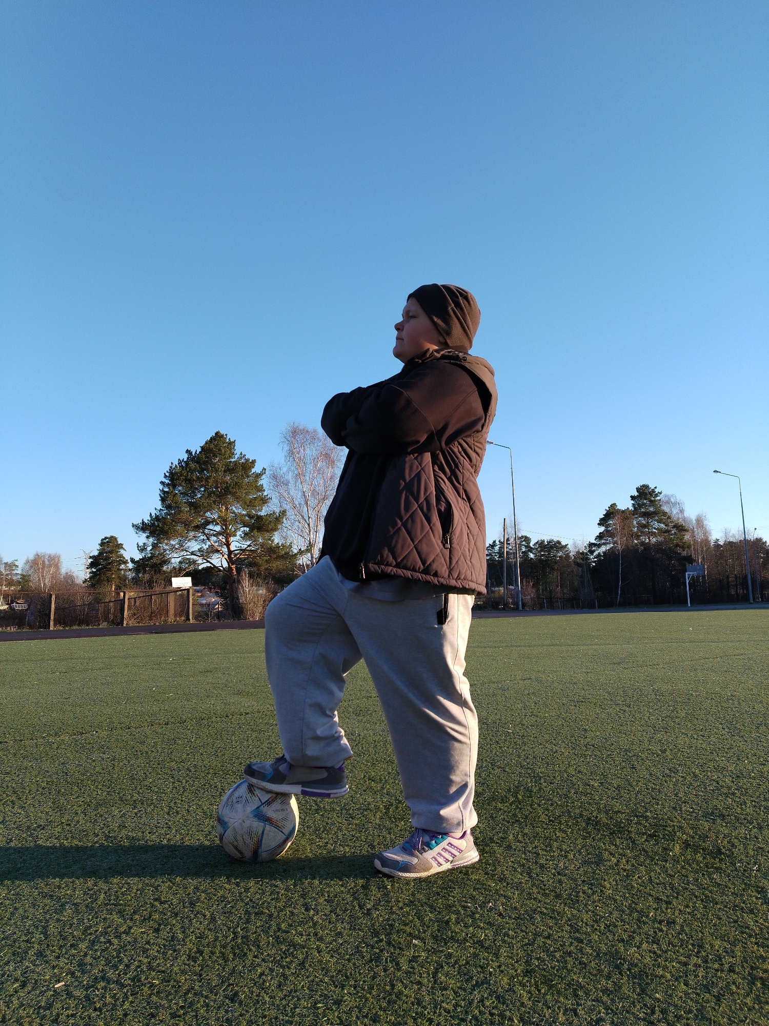 A full body shot shows a young person standing on a green sports field, appearing to be in Chaikovsky, Russia. They are standing with one foot on a soccer ball. The person is wearing light gray sweatpants, and multicolored sneakers, a brown quilted jacket, and a brown beanie. They have their arms crossed and are looking to the side. Behind them is a fence, a row of trees, and lampposts. The sky is clear and blue. The overall impression is of a casual moment captured outdoors on a sunny day.