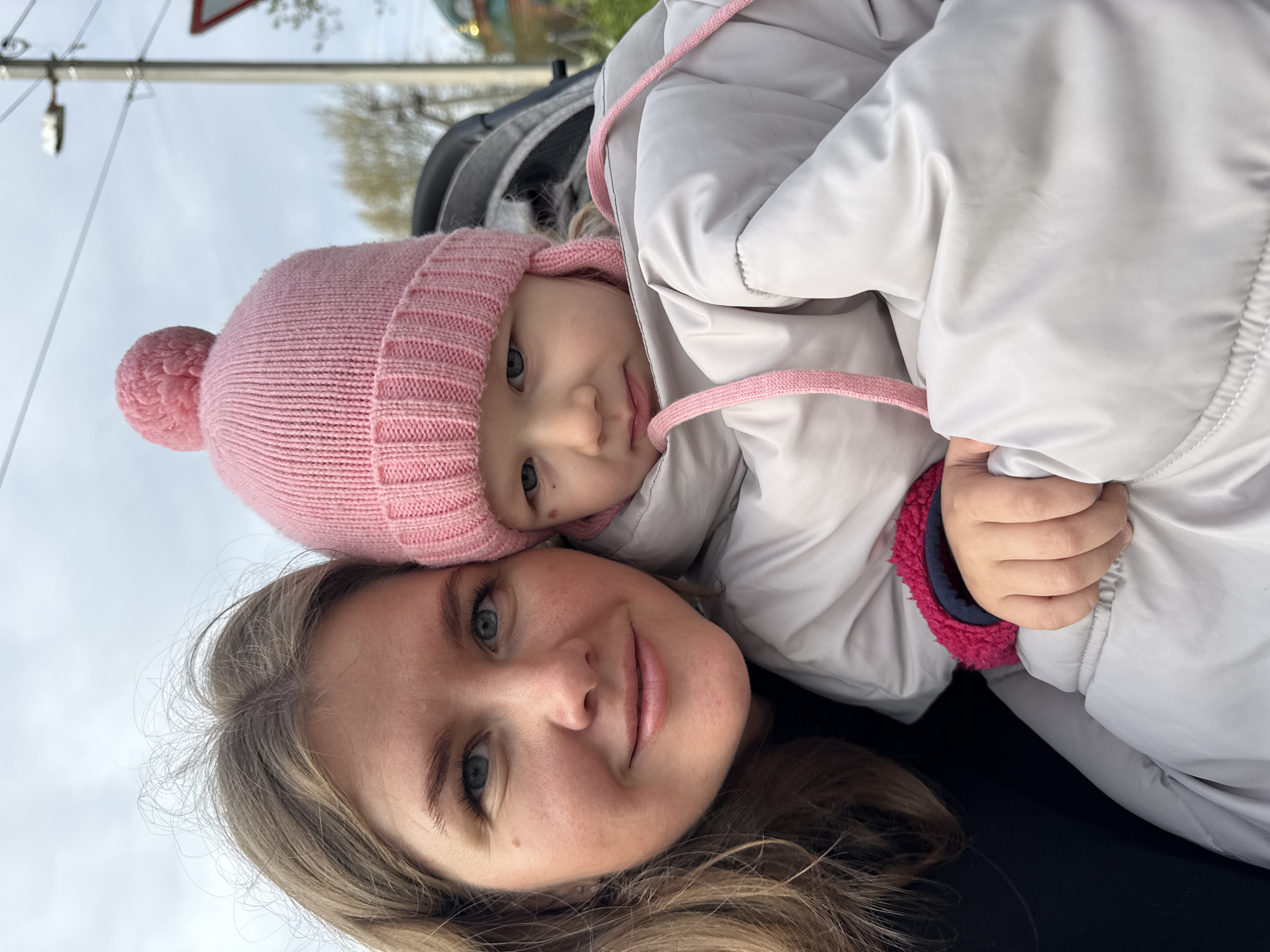 A woman and a young child are pictured outdoors, possibly in or near the city of Ферма, Russia. The woman, with light brown hair and fair skin, is smiling at the camera. The child, wearing a pink knit hat, is nestled against the woman, appearing to be held by the woman's right hand. The child is also wearing a light-colored jacket with pink trim. The background includes a street sign and glimpses of other buildings and trees.