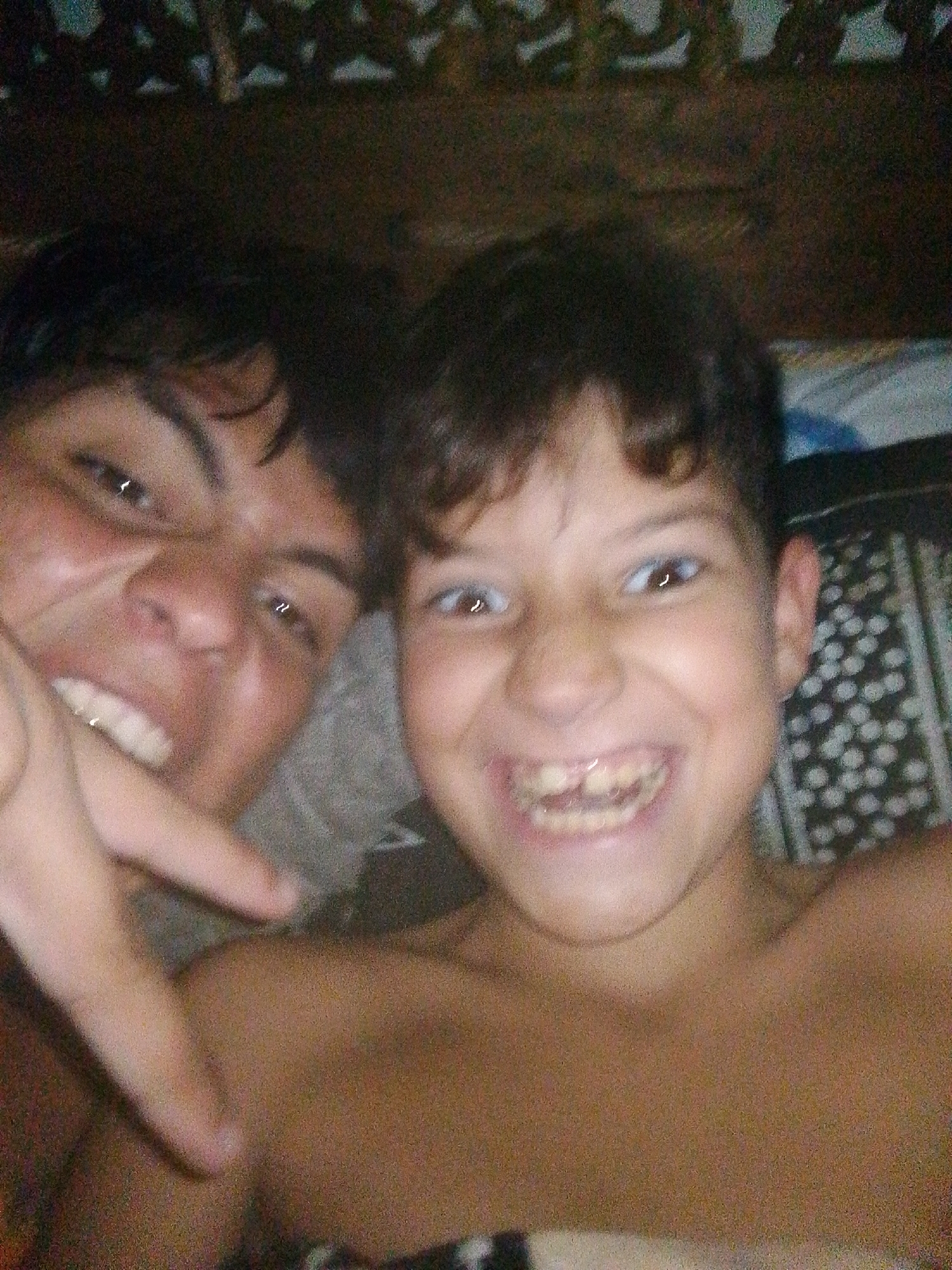 The image is a close-up, slightly blurry selfie of two young boys. The boy in the foreground on the left is looking directly at the camera with a wide, toothy grin, revealing some missing teeth. He is topless, with his left arm extended towards the camera, his hand partially obscuring his face. His skin appears tanned, and his hair is dark and somewhat messy.
The boy to the right, also topless, is slightly further back and to the side, his face also visible in the frame. He is also smiling, though less prominently than the boy on the left. Both boys appear to be in an indoor setting, possibly a bedroom, with a patterned blanket or sheet visible behind them. The lighting suggests it could be daytime, but there are no clear indicators of the time of day or weather. The overall emotion conveyed is one of playfulness and camaraderie. No text is visible in the image.