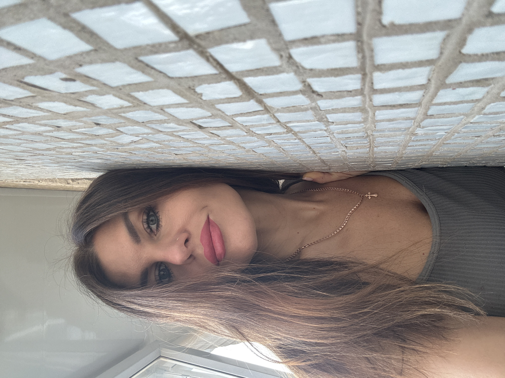 A woman with long brown hair, light skin, and green eyes is posing indoors. She wears a gray tank top and a gold necklace with a cross pendant. The background shows a wall or ceiling covered in small, white, square tiles. The setting could be Chaikovsky, Russia, but there are no identifying characteristics in the image to confirm this. The woman is positioned at an angle, with the tiling appearing directly above her. A window is visible in the lower left corner.