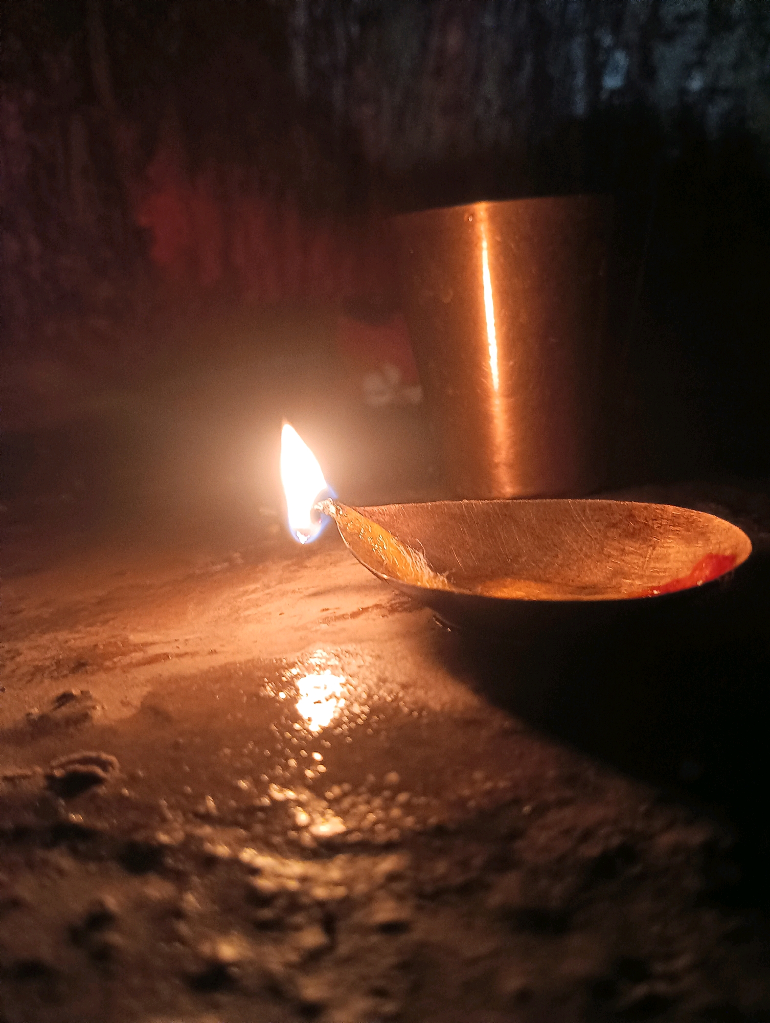 In an indoor setting with low lighting, a close-up shot focuses on a lit diya, a traditional Indian oil lamp. The diya, made of brass, sits on a rough, wet surface that reflects the warm glow of the flame. The flame itself is bright orange and yellow, with a hint of blue at its base, casting an ethereal light. In the background, a copper pot is visible, also illuminated by the diya's light. The overall mood is serene and atmospheric, suggesting a moment of quiet contemplation or prayer. The scene appears to be set on the floor of a room, with the rough texture of the surface indicating it might be an unpolished stone or concrete floor. The water on the surface adds to the reflective quality of the scene, creating a mirror-like effect for the diya's light.