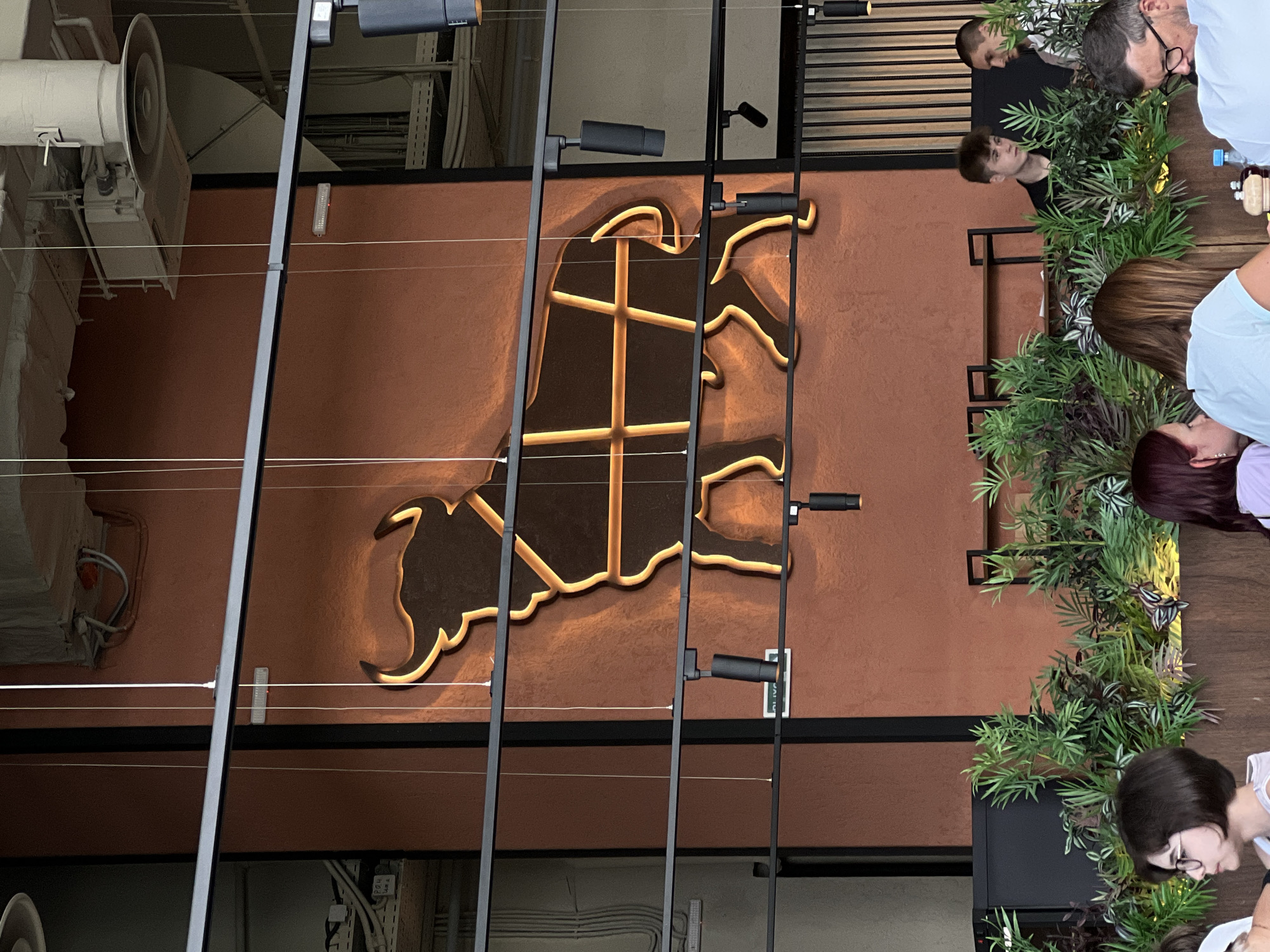 The image shows a group of people seated around a table in what appears to be an indoor dining establishment. The table is adorned with lush green plants, creating a naturalistic ambiance. The focal point on the wall behind them is a large, stylized silhouette of a bull, illuminated with a warm, golden light, suggesting it's a logo or decorative element.
Several individuals are visible, their faces partially obscured or turned away from the camera. One person on the right side of the frame, wearing a white top, is looking down at the table. Another person, wearing glasses, is also engaged with something on the table. In the background, a young man with short hair is looking towards the left. There is also a glimpse of another person behind him. The people appear to be casually dressed, and the overall atmosphere seems relaxed.
The lighting suggests it could be daytime, or an indoor space with artificial lighting. The scene is likely a restaurant or cafe, given the table setting and the presence of multiple diners. The bull motif on the wall hints at a potential theme, possibly related to meat, power, or a specific brand identity. There are no explicit cues regarding the time of day or weather outside. The location is identified as Khimki, Russia. No discernible text is visible in the image.