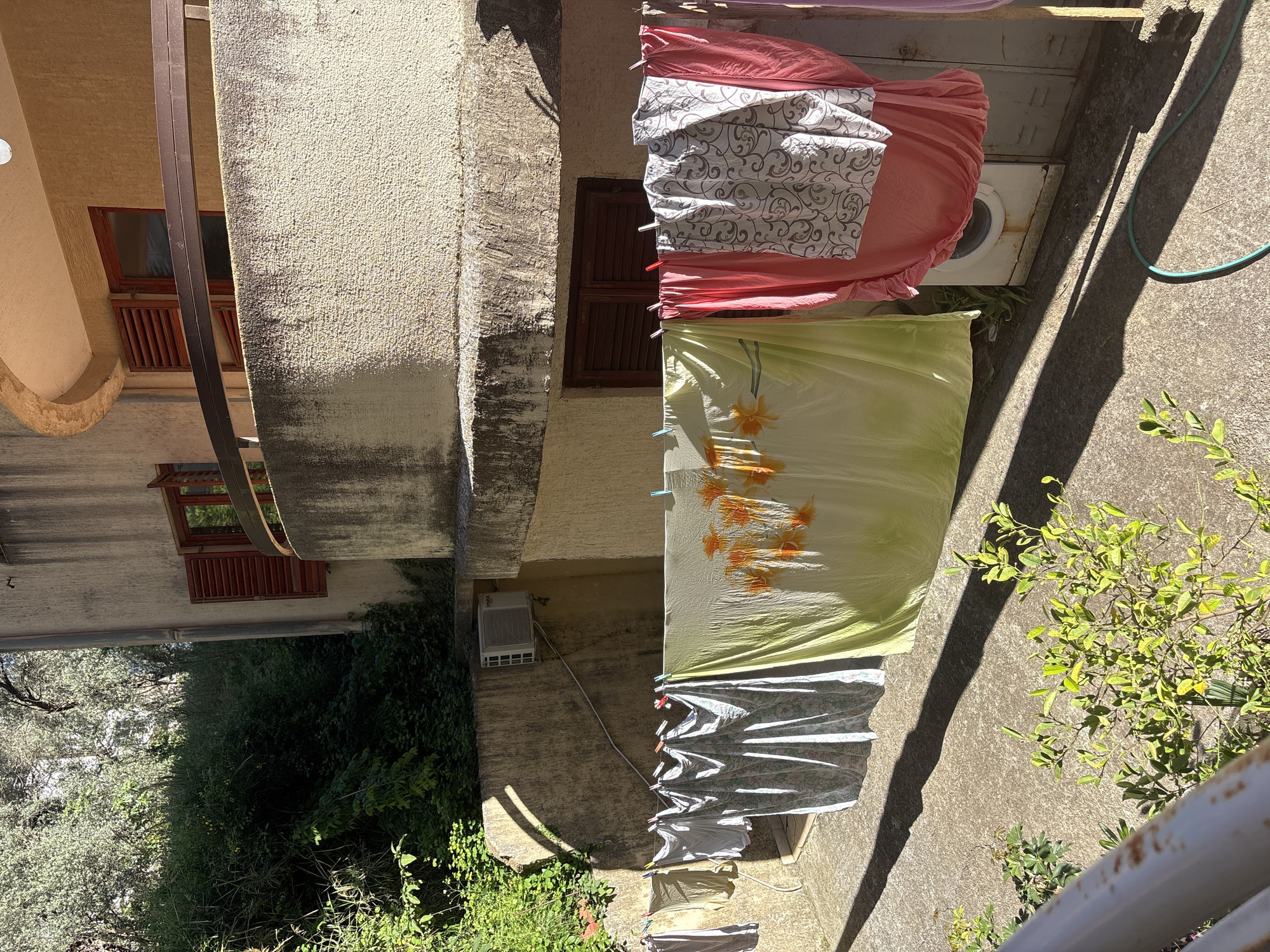 The image shows an exterior courtyard or balcony area in Bečići, Montenegro. A line of laundry hangs in the foreground, including a pink sheet, a white patterned sheet, and a yellow floral patterned sheet. A white washing machine sits on the ground. Adjacent to the laundry is a concrete surface with plants and a green garden hose. Buildings surround the area, featuring tan stucco and windows with wooden shutters. Trees and greenery fill the lower left corner of the image.