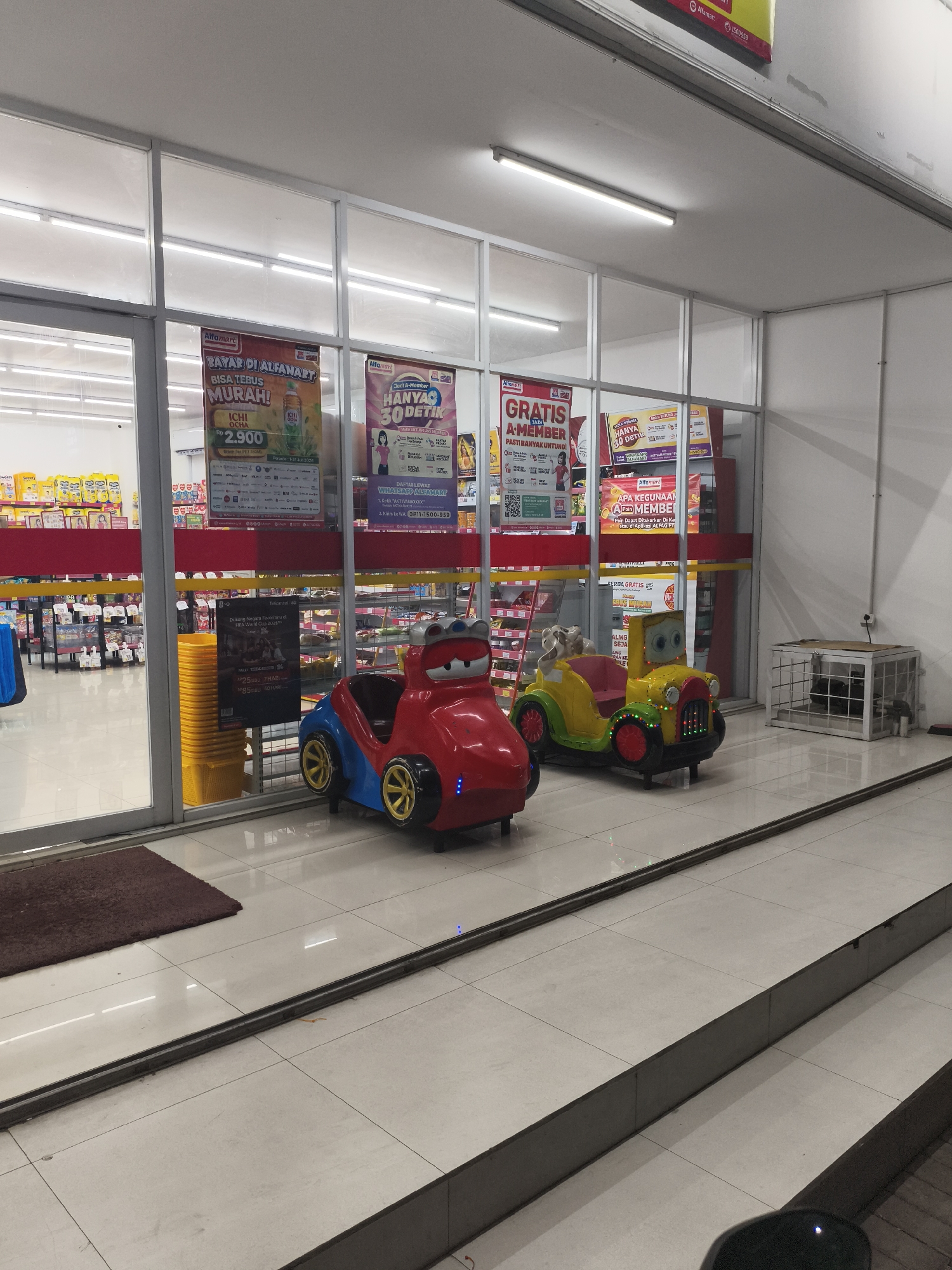 This is an exterior shot of a minimarket in Depok, Indonesia, likely a branch of Alfamart, indicated by signage. The storefront features a large glass facade with a red trim. Inside, shelves stocked with various products are visible. In front of the store, two coin-operated amusement rides shaped like cartoon cars are positioned on a paved surface. A red car-shaped ride with blue accents is to the left, and a yellow car-shaped ride with a green hood and red wheels is to the right. A dark brown mat is placed on the ground to the left of the rides. A stack of yellow baskets is visible just inside the entrance to the right of the mat. A caged structure containing what appears to be machinery or equipment is situated further to the right. The lighting suggests it is either evening or a dimly lit interior. The ground is tiled with light-colored square tiles. A metal track runs along the ground in front of the rides and the minimarket entrance.