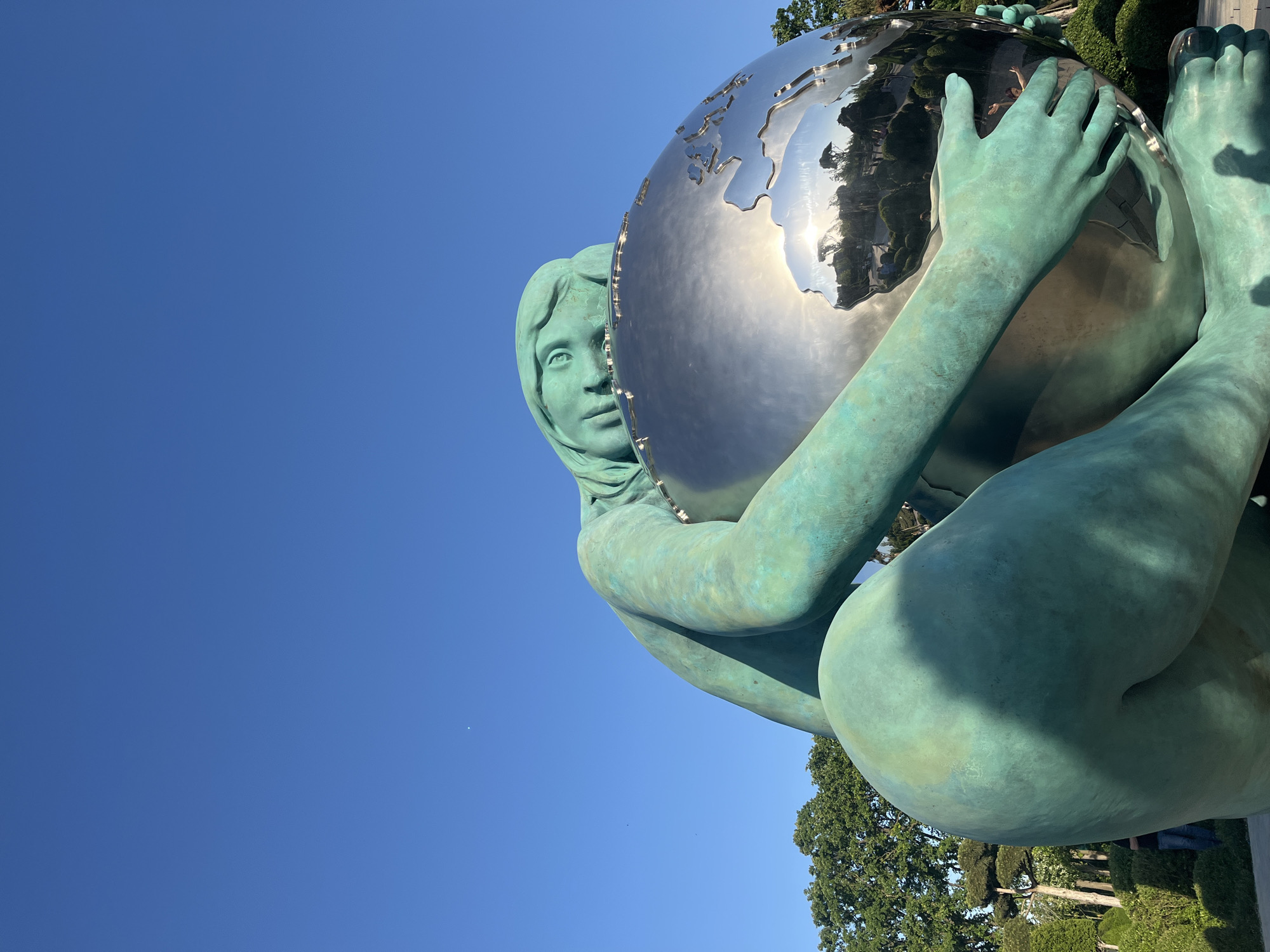 The image shows a close-up, low-angle view of the "Girl Holding the World" sculpture in Krasnodar, Russia. The sculpture depicts a female figure with a verdigris-toned body and face, cradling a polished metal globe representing the earth. Her hands and feet are clearly visible as she embraces the globe. The globe's surface reflects the surrounding environment, including trees and the sky. The background features a clear blue sky occupying a significant portion of the upper left, and greenery representing trees and bushes toward the lower right. The sculpture appears to be positioned outdoors within a park-like setting.
