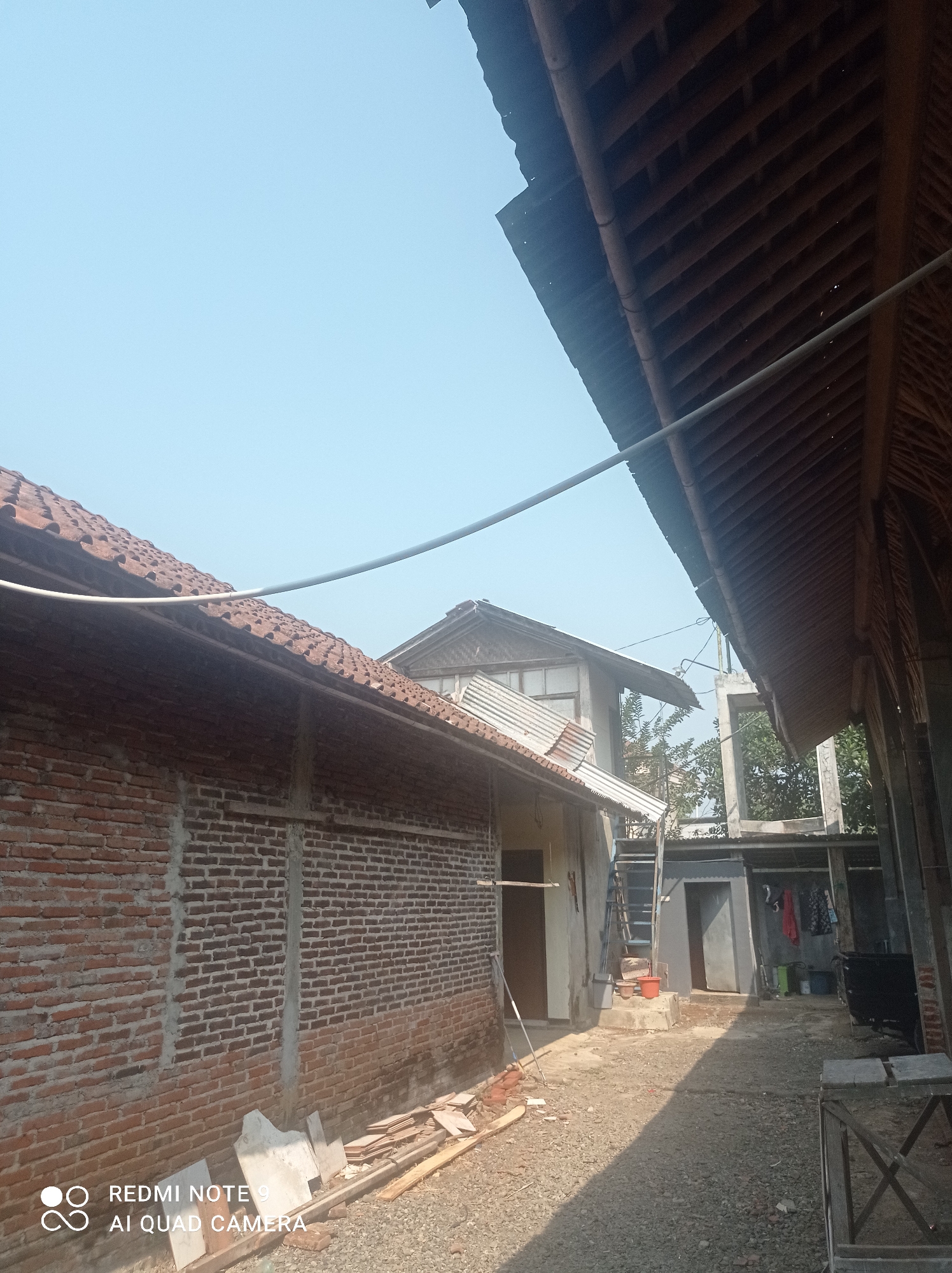 The image shows a courtyard in a semi-developed area. The foreground features a gravel ground with scattered building materials, including planks of wood and what appear to be concrete slabs. To the left, a brick wall of a building is visible, extending into the middle ground. Above the brick wall, a tiled roofline is prominent. In the center of the image, a light-colored building with a corrugated metal roof is present, showing construction elements like a ladder leaning against it. Further back, an unfinished concrete structure with an open frame and trees behind it can be seen. Various objects are scattered in the courtyard, including buckets, clothing hanging on a line, and some containers. The sky above is clear and light blue. The scene suggests ongoing construction or renovation work in a residential or semi-industrial area within the city of Cipawon, Indonesia.