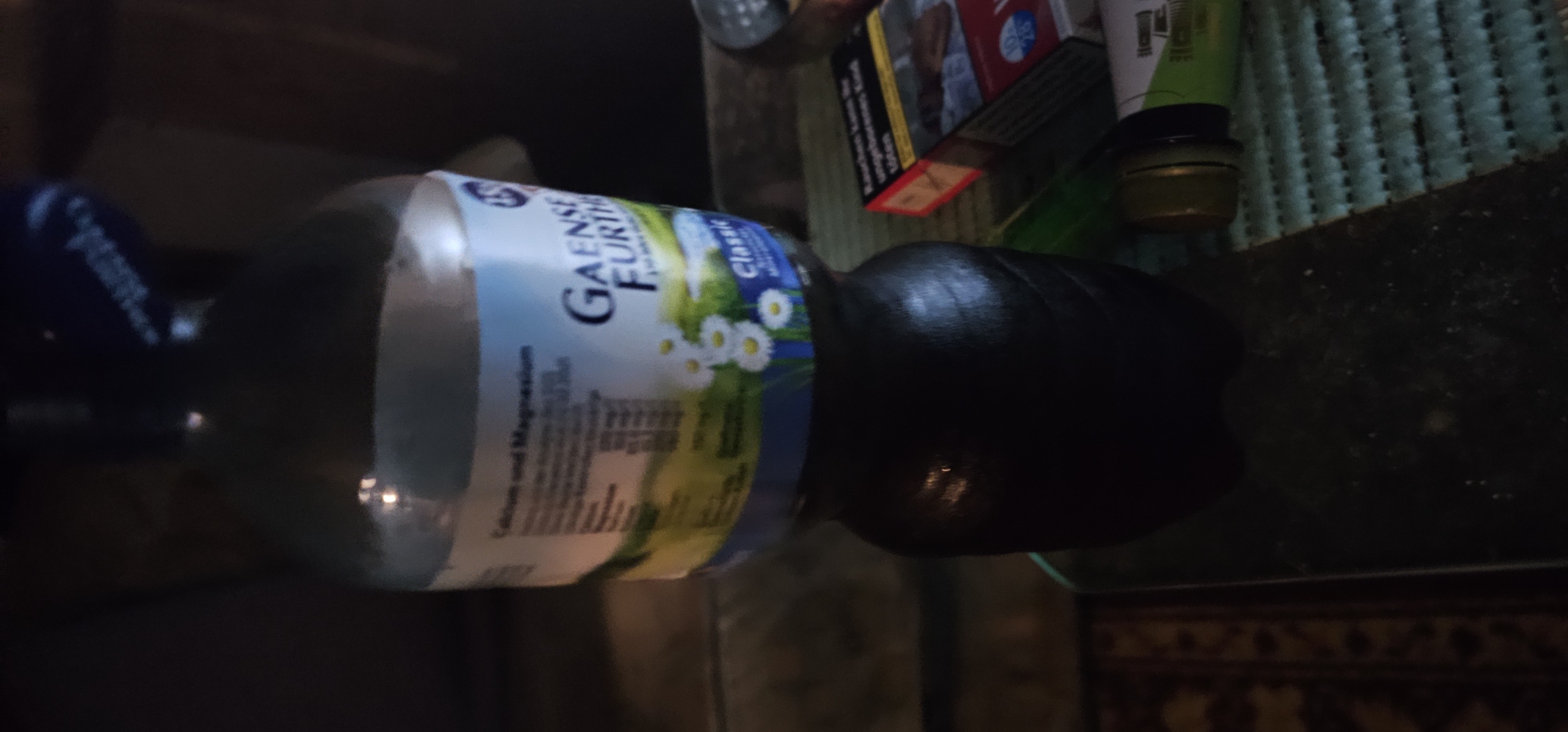 A dark-colored liquid is contained within a transparent bottle, which rests on a table or flat surface. The bottle has a label with text and images, including white daisies against a blue and green background. Part of the label reads "Calcium und Magnesium" and "Gaense Further Classic". To the right of the bottle, a pack of cigarettes with a red and yellow design is visible. A portion of another object, possibly a bottle or container with a green label and a gold cap, is also present in the foreground. The background is dark and out of focus, with some suggestion of indoor lighting. The scene appears to be an indoor setting, possibly a room in Schönebeck (Elbe), Germany, given the context. There are no individuals directly depicted in the image.
