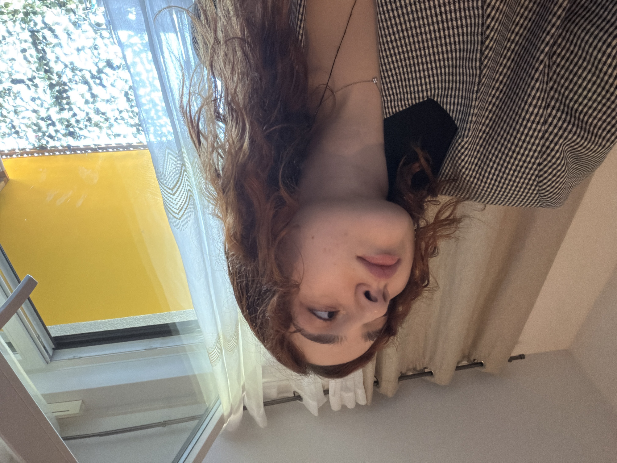 This media file captures a close-up of a young woman in an indoor setting in Strasbourg, France, presented in an upside-down orientation. The woman, with shoulder-length, wavy, reddish-brown hair, occupies the central left portion of the frame. She has fair skin and a delicate necklace with a small, square-shaped pendant. She is wearing a black and white plaid jacket over a black garment. Her eyes are cast slightly to her left (viewer's right), and her lips are slightly parted, giving her a pensive or contemplative expression.
The background features a window on the left side, through which bright daylight streams. Outside the window, a large, vibrant yellow surface is visible, likely an exterior wall or awning, reflecting intense sunlight. Above the yellow, abundant green foliage is discernible, suggesting trees or bushes under a bright sky. White, sheer curtains with a subtle geometric pattern are drawn across part of the window, diffusing the sunlight. To the right, a solid beige curtain hangs from a rod, partially obscuring the background. The interior walls appear to be a light, neutral color. The bright lighting suggests it is daytime, likely a sunny day. No visible text is present in the image.