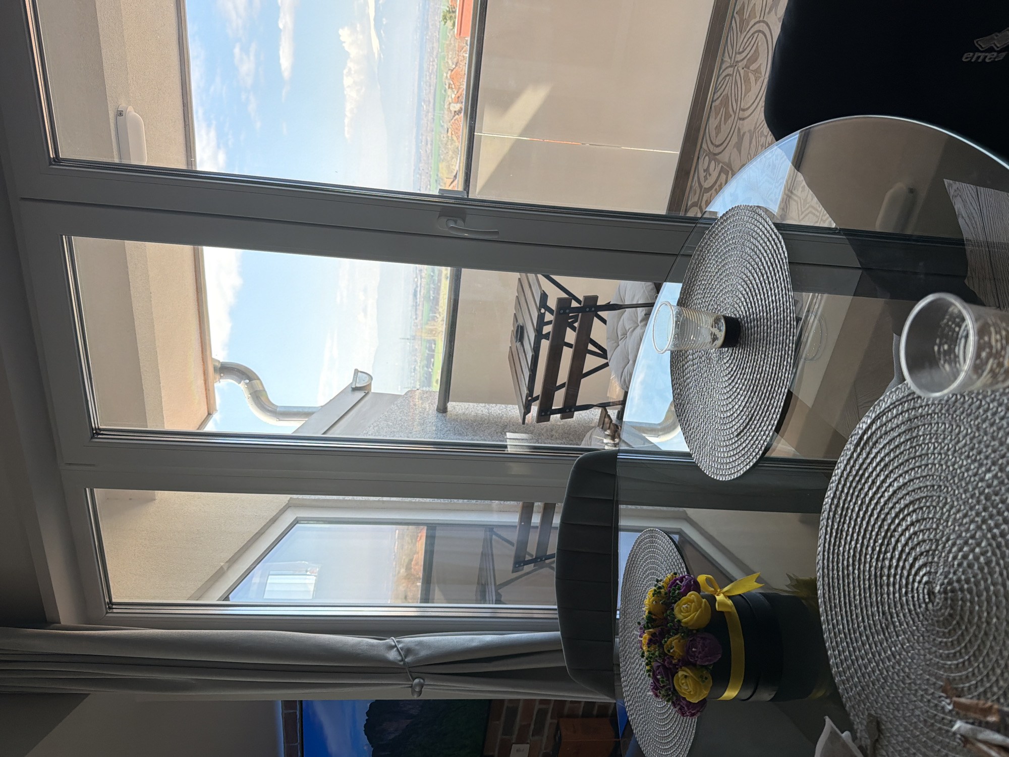 A partial interior view shows a clear glass table with woven placemats and glasses. A floral arrangement in a black cylindrical box with a yellow ribbon sits on a placemat. The room opens to a balcony with a table and chairs, beyond which is a cityscape under a blue sky with scattered clouds. The location is suggested to be Buhovo, Bulgaria, due to the prompt.