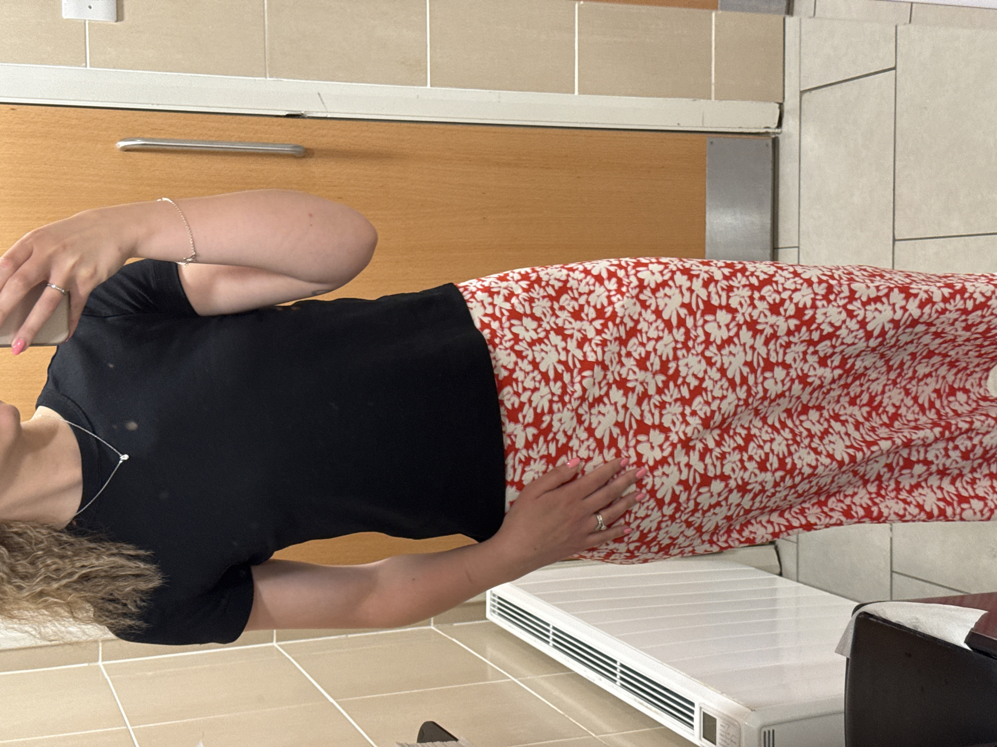 A woman is seen indoors, likely within a residential space, possibly in Ipswich, United Kingdom, given the assumption based on the prompt. She is wearing a black short-sleeved top and a long, red and white floral print skirt. Her hair appears to be blonde and curly. She has light skin tone. Her hands are visible, adorned with rings, and her fingernails are painted pink. The background consists of a kitchen with light brown cabinetry and a tile wall. A white radiator or appliance is visible near her skirt. No specific actions or interactions are evident from the image, only her presence in the space.
