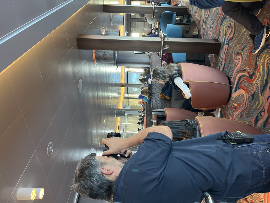 A man with graying hair is taking a picture with a camera in a room with a crowd of people. He's wearing a navy blue shirt and jeans, with a walkie-talkie clipped to his waistband. The room has unique, rounded chairs with pinkish-brown upholstery and a colorful patterned carpet. A vertical structural element with a decorative stone detail is visible. People are seated, some facing a screen or window.