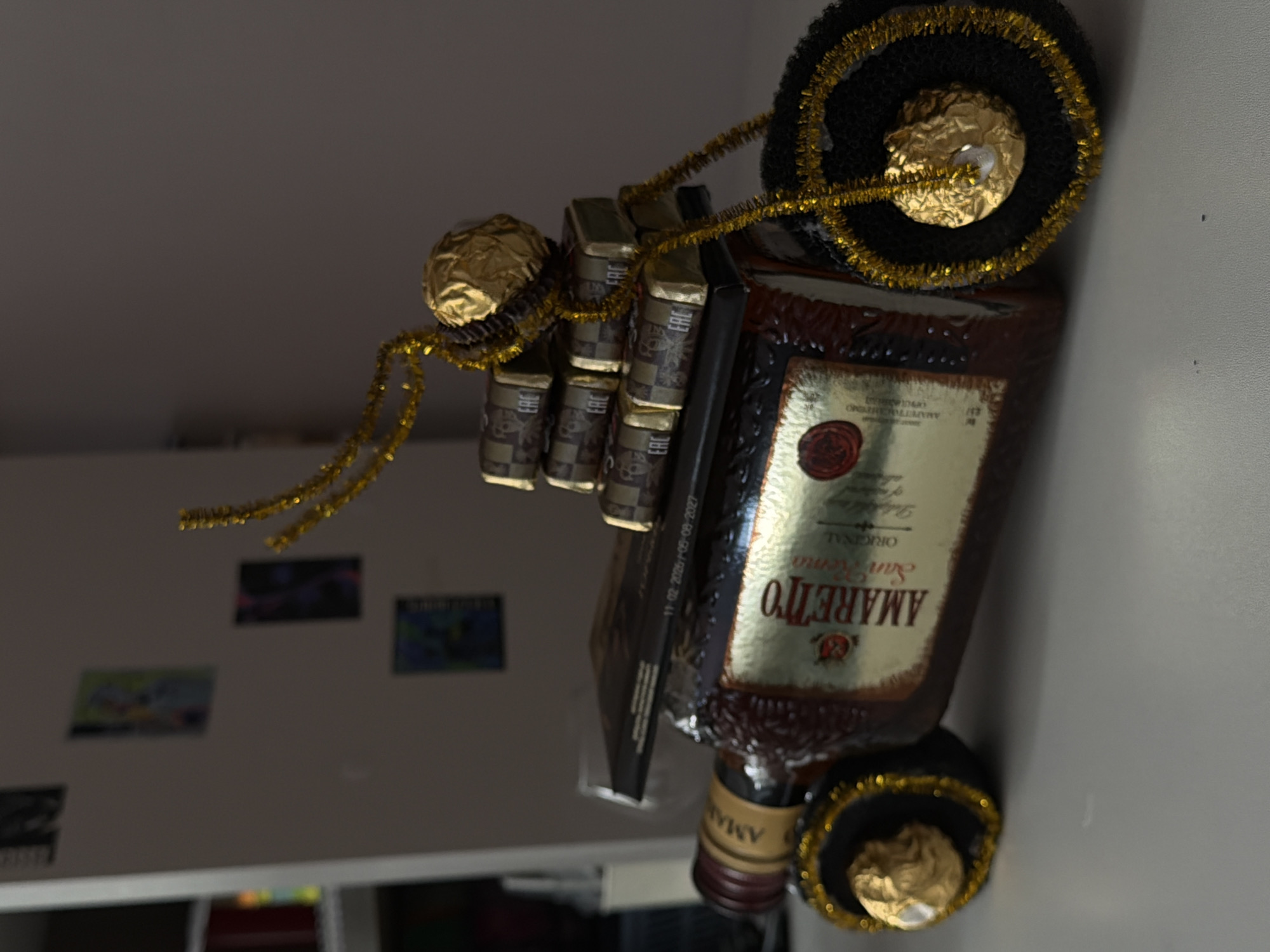 The image displays a decorated dark brown glass bottle of Amaretto liqueur, positioned horizontally, suggesting it is part of a gift or a festive display. The bottle's main label clearly shows "AMARETTO ORIGINAL SAN REMO" in gold script on a golden background, along with a red wax-seal emblem and the word "LIQUORE."
The bottle is intricately adorned with multiple gold-wrapped chocolate candies, resembling Ferrero Rocher, and several smaller, dark-wrapped rectangular items, likely mini chocolate bars or similar treats. These decorations are secured with strands of shiny gold tinsel or pipe cleaners, which also wrap around the neck and body of the bottle. On some of the dark-wrapped items, the "EAC" (Eurasian Conformity) mark is visible, indicating compliance with regulations for products sold within the Eurasian Customs Union, which includes Russia. A black strip attached to the bottle displays dates: "11-02-2020" and "09-09-2027," likely indicating production and expiry.
At both ends of the bottle, there are black, circular, textured pads, possibly coasters or decorative elements, each containing a gold-wrapped chocolate in its center, also framed by gold tinsel.
The setting appears to be an indoor space, likely a home, with a light-colored wall in the background. Several small, blurred rectangular pictures or prints are affixed to this wall, one faintly showing a cartoon character. A horizontal white ledge or surface is visible towards the bottom of the background. The lighting is bright and even, consistent with indoor illumination. No people are visible in the image. The overall arrangement evokes a sense of celebration or gifting.