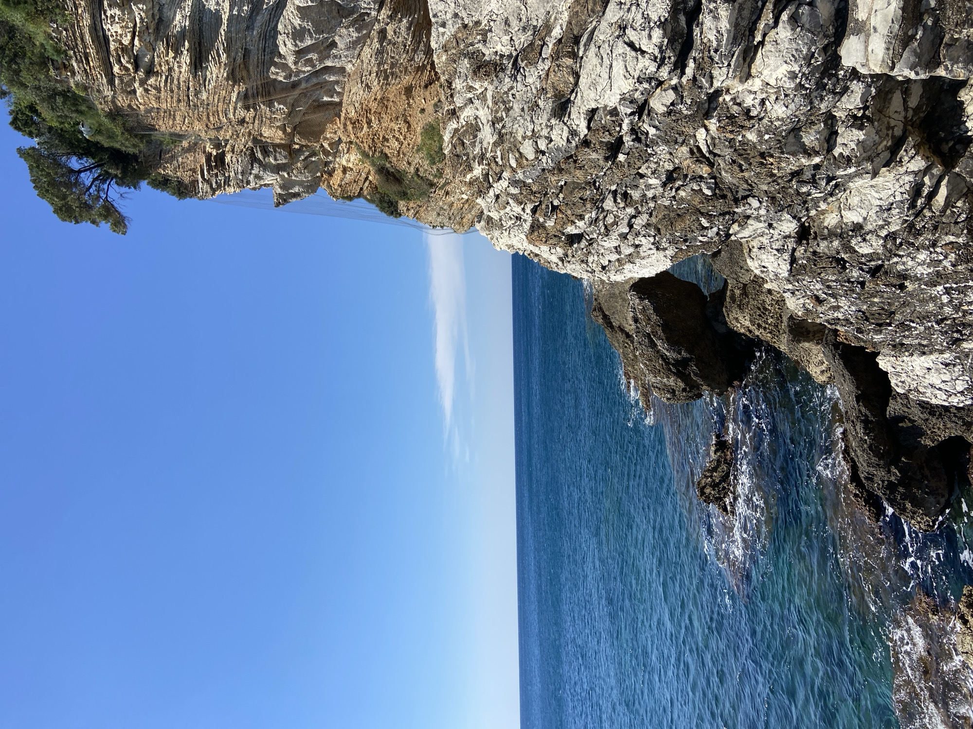 The image depicts a coastal scene in Budva, Montenegro, showing a rocky cliff formation meeting the Adriatic Sea. The cliff on the right is textured, with visible rocks and water washing at the base. On the left, the cliff appears to be layered with sparse vegetation on top. A netting system is visible along the cliff to prevent rocks from falling. The sea stretches out, transitioning from a darker hue near the cliffs to a lighter blue towards the horizon where it meets the sky. The sky is a clear, bright blue with a faint cloud streak in the center.