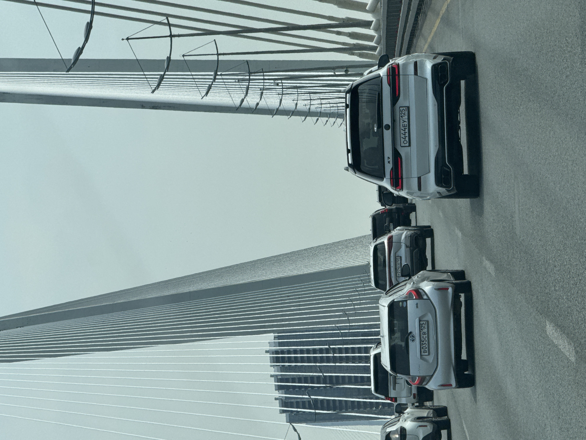 The image depicts a line of cars traveling on the Russky Bridge in Vladivostok, Russia. The cars are primarily silver or white, and their license plates are visible. The bridge's suspension cables and support structure are prominent in the background, and a tall building or structure can be seen reflected in the bridge's surface. The weather appears overcast, with a grey sky. The road surface is asphalt, and lane markings are visible.