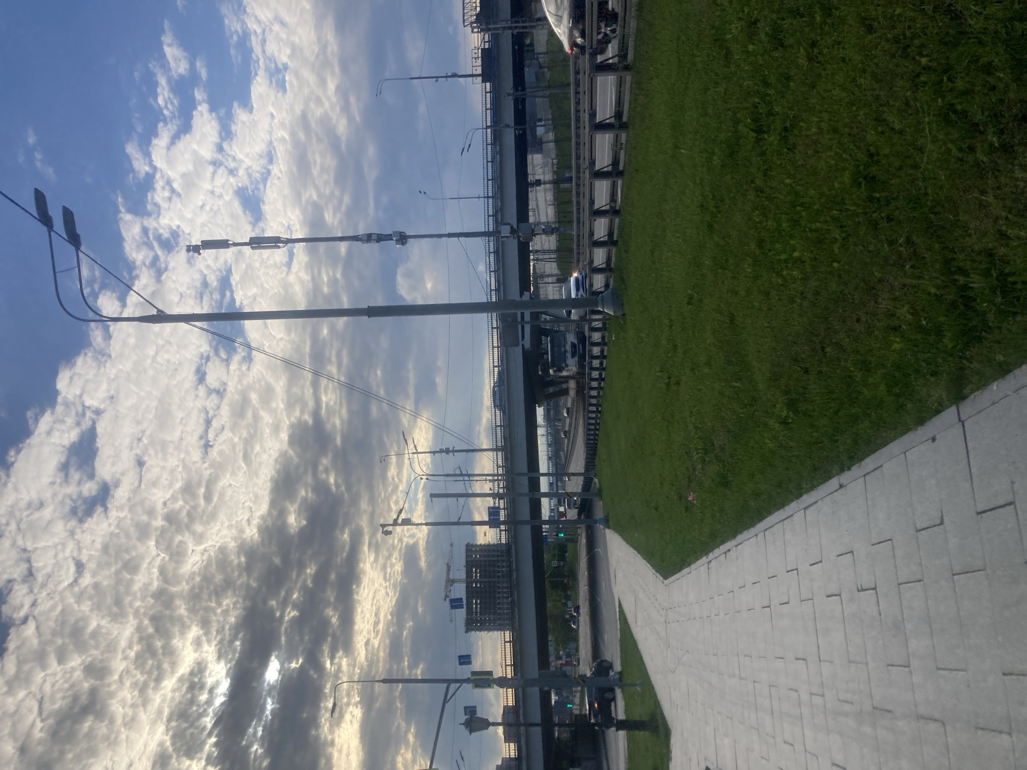 The image depicts a pedestrian walkway in Moscow, Russia, bordering a grassy area. Above, a highway or bridge is visible, with vehicle traffic moving across it. The sky is filled with clouds, and several streetlights line the walkway. In the background, there is a building under construction.