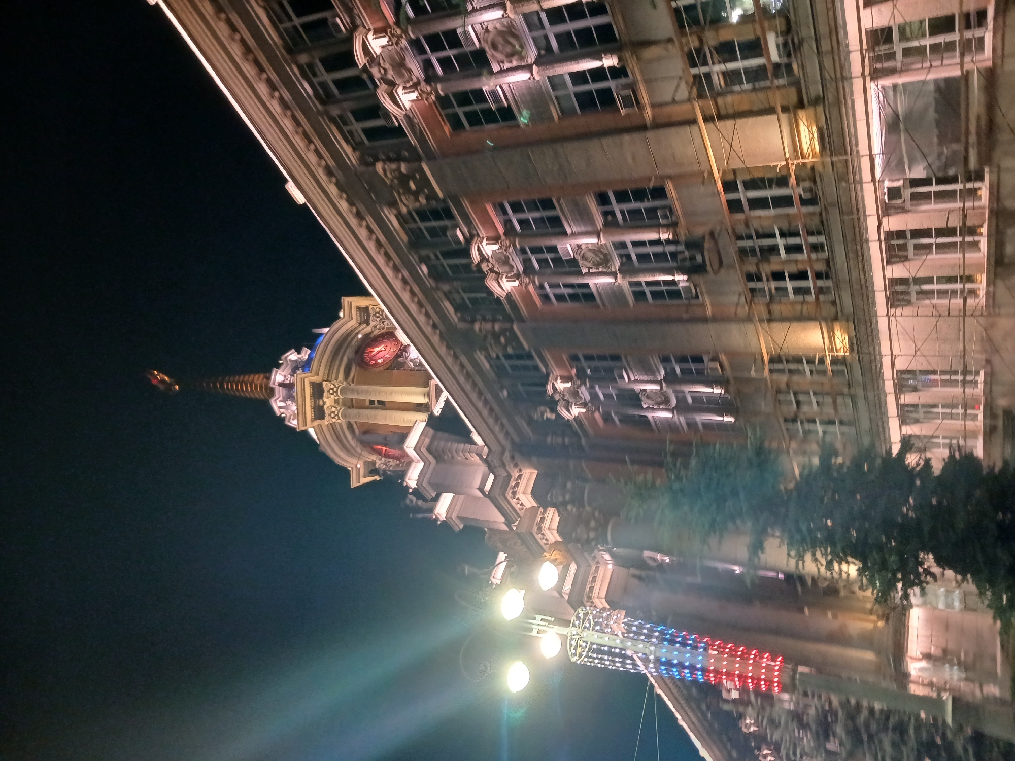 The image captures a nighttime view of the Sverdlovsk Oblast Government Building, also known as the White House, located in Yekaterinburg, Russia. The building's facade is illuminated by warm, artificial lights, highlighting its architectural details. A prominent spire crowns the central section of the building. To the left, a street lamp with multiple globes is visible, adorned with a festive string of lights in red, white, and blue, suggesting a celebration or holiday. A tall, dark evergreen tree stands in the foreground, partially obscuring the building. Scaffolding is present on some sections of the building, indicating ongoing construction or renovation work. The sky is dark, typical of nighttime. No people are visible in the frame.