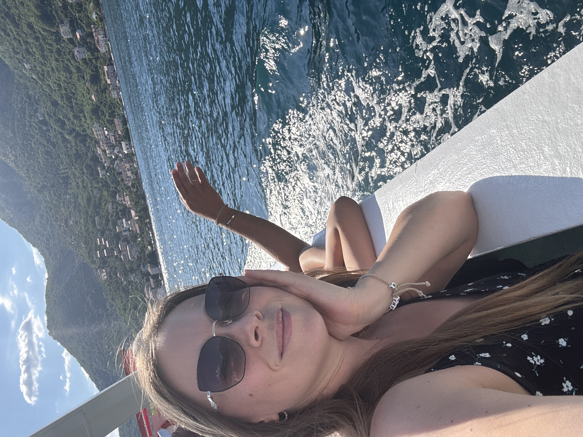 The image depicts a young woman with long brown hair and sunglasses, leaning on a boat railing. She is looking towards the camera with her hand resting on her cheek. Behind her, a second person's arm is visible, extended outward, possibly waving. The boat is in motion, creating a wake in the deep blue water. In the background, lush green hills with scattered buildings rise steeply from the coastline, characteristic of Lepetane, Montenegro. The sky is blue with scattered clouds, and the sun is casting bright reflections on the water.