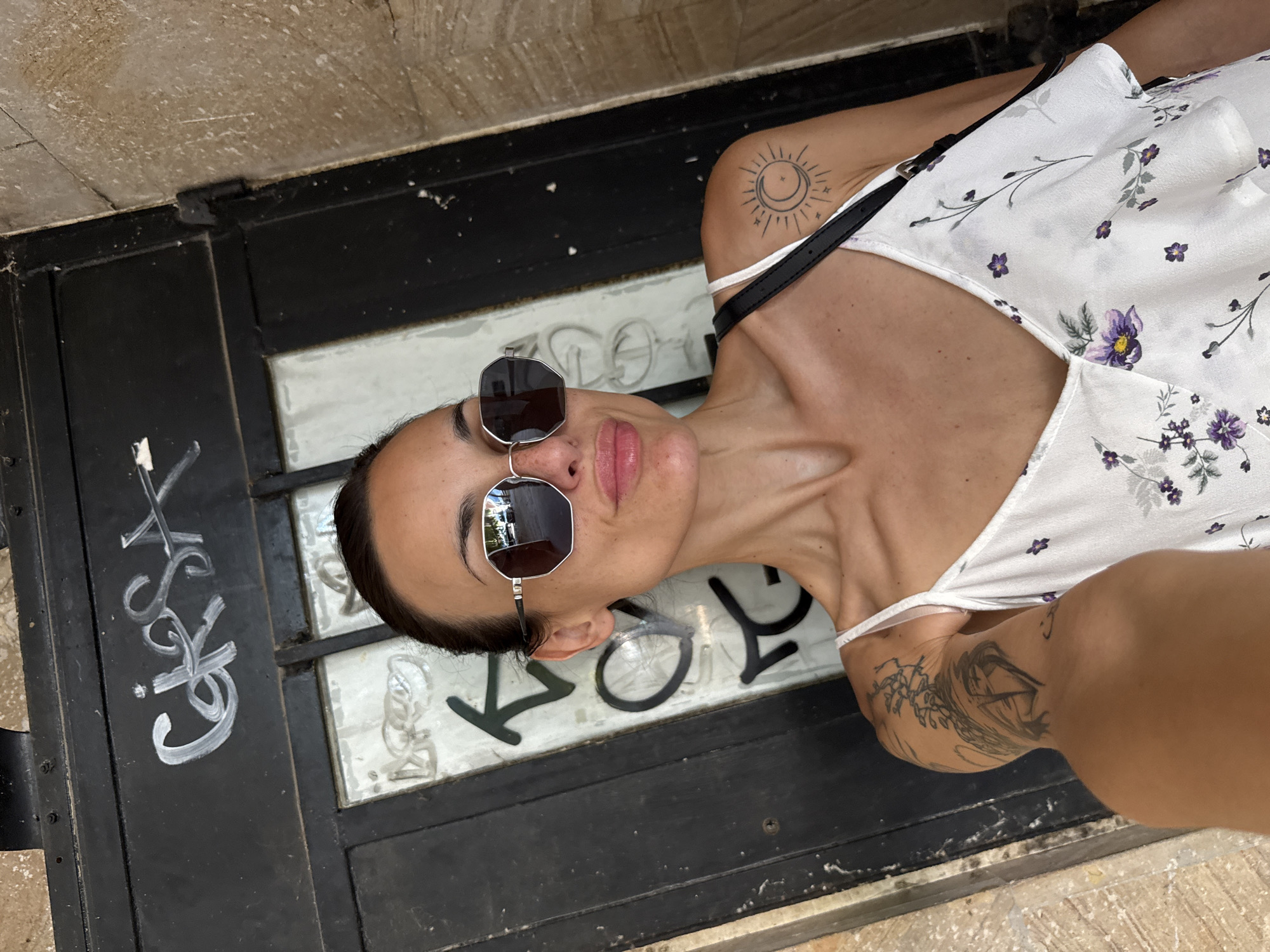 A woman with dark hair pulled back is taking a selfie in front of a dark metal door with graffiti. She is wearing large, octagonal sunglasses and a white camisole with a floral print. She has tattoos on her right shoulder and arm, including a sun and moon design on her shoulder. The door is marked with white graffiti, and one of the windows on the door reflects a street scene with cars. The building has a rough, stone facade. The image appears to have been taken outdoors in a city.