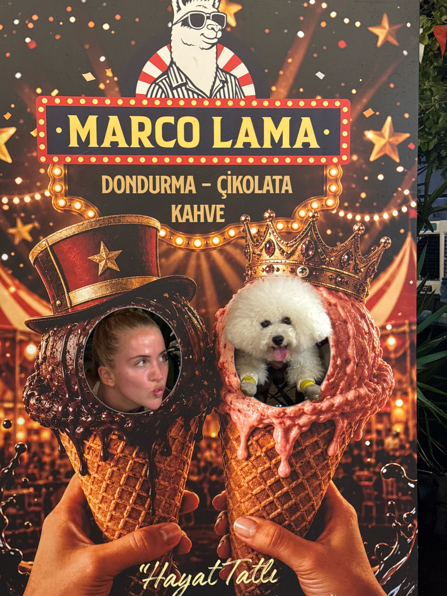 This is a photograph of a promotional poster for "MARCO LAMA," an establishment that appears to sell "DONDURMA - ÇIKOLATA KAHVE," which translates from Turkish to "Ice Cream - Chocolate Coffee." The poster features a stylized llama wearing sunglasses and a striped shirt at the top, framed by decorative lights and stars. The main focus of the poster is two large ice cream cones, each with a circular cutout where faces are inserted. In the left cone, a young woman with blonde hair pulled back is shown with her mouth puckered, looking to the right. She is positioned within a dark brown, chocolate-flavored ice cream cone topped with dripping chocolate, and wearing a top hat with a star. In the right cone, a fluffy white Bichon Frise dog is visible, wearing a golden crown and small yellow and grey booties, peeking out from a pink ice cream cone. The bottom of the poster displays the text "Hayat Tatlı" which means "Life is Sweet" in Turkish, and the phrase "Antalya Turkey" is present at the bottom. The overall aesthetic is festive and playful, using a bright and saturated color palette.