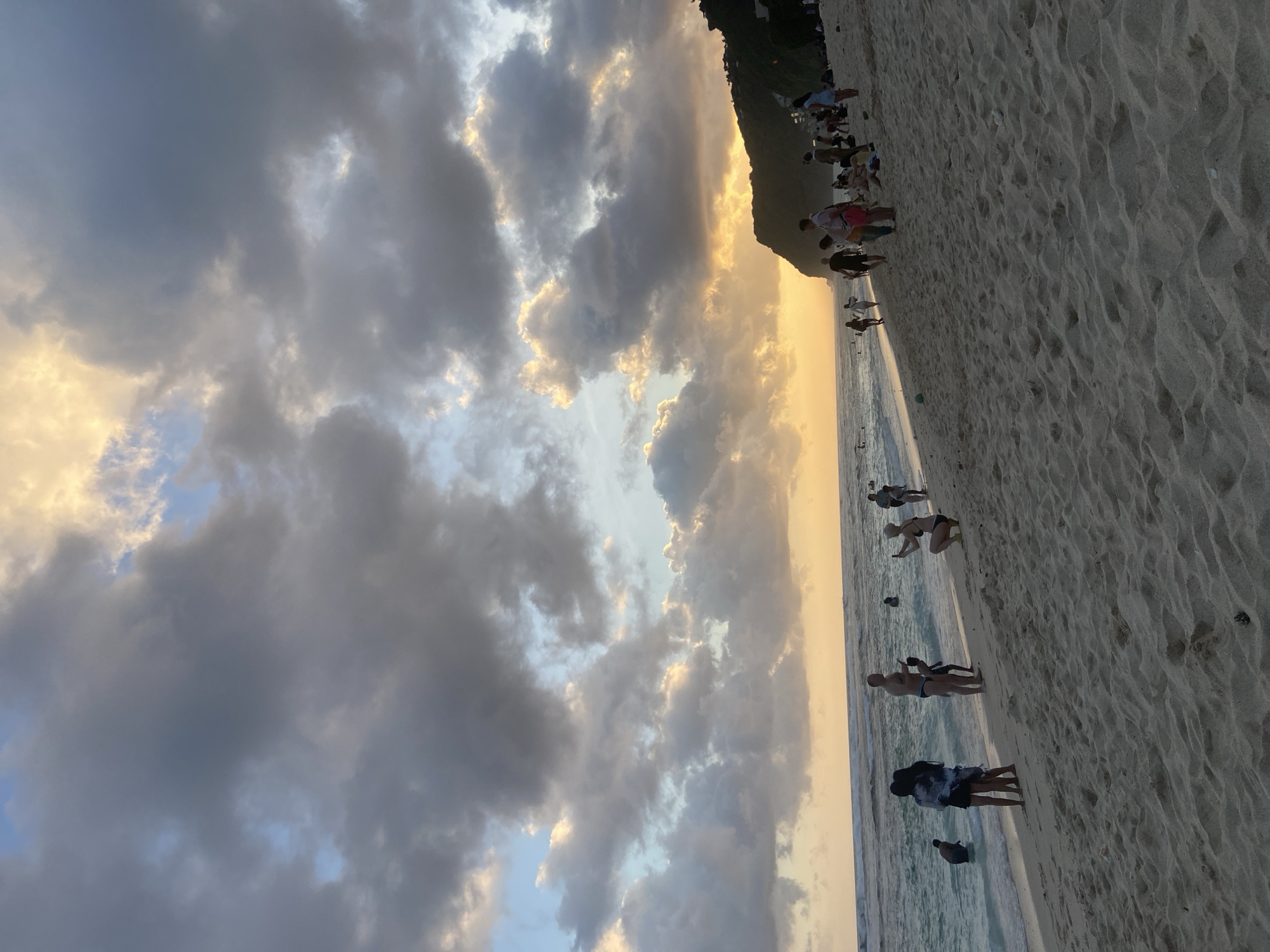 The image depicts a beach scene in Kuta Selatan, Indonesia, during what appears to be sunset. The sky is filled with dramatic clouds, tinged with orange hues from the setting sun. The beach is sandy and stretches into the distance. People are scattered along the shoreline, some standing, others walking, and some appearing to be swimming or wading in the water. A tree-covered hillside runs along the back of the beach.