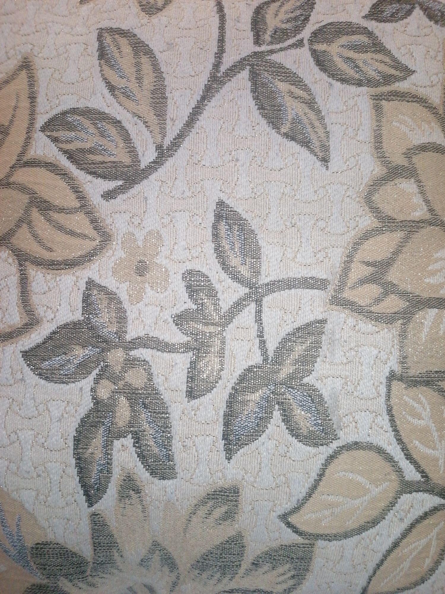 The image shows a close-up view of a fabric with a floral pattern. The pattern consists of stylized leaves and a small flower, rendered in shades of beige, cream, and grey with a subtle metallic sheen. The background fabric is textured and appears to be a light cream or off-white color. There are no people or any indication of a specific location, either within Angarsk, Russia, or elsewhere. The image is a purely visual representation of a textile design.