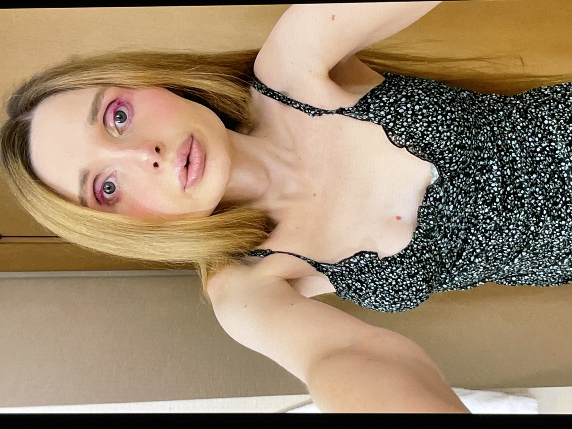 A woman with blonde hair and dramatic pink eyeshadow is shown in a close-up, angled shot. She is wearing a black dress with a small white floral pattern and a scalloped neckline. Her head is tilted, and her gaze is directed towards the camera. Her lips are slightly parted, and she has rosy blush on her cheeks. The background appears to be a plain, neutral-colored surface. The lighting suggests an indoor setting, possibly with artificial light. No text is visible in the image.