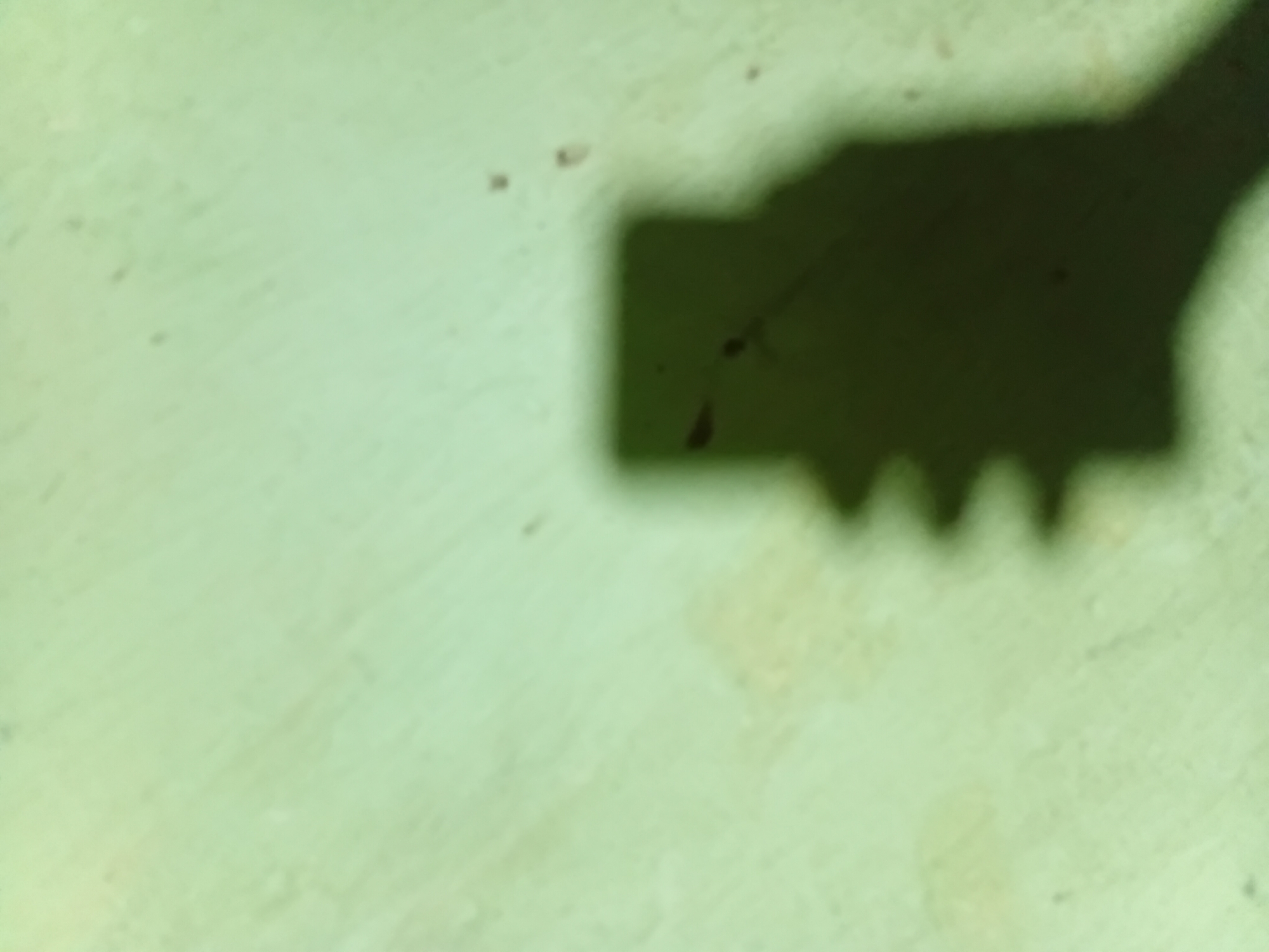 The image shows a shadow of a person holding a smartphone, casting a silhouette on a light green, textured wall. The shadow is dark and clearly defines the outline of a head and hand holding a rectangular object, likely the phone. There are a few small, dark spots on the wall, possibly dust or minor imperfections. The lighting suggests an indoor setting, with the light source positioned to create this prominent shadow. There is no specific information about the location in Nkpor, Nigeria, other than it is a wall within that city.