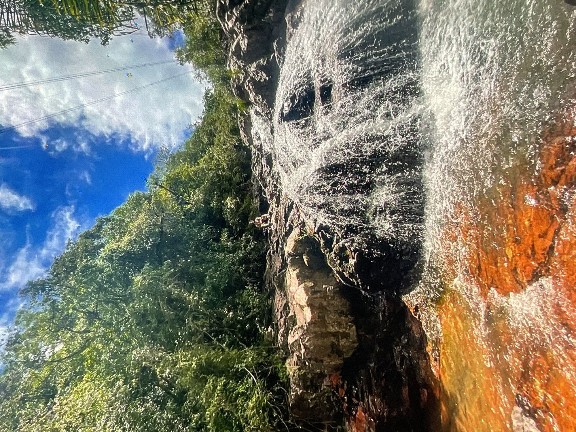 The image depicts a waterfall flowing down a rocky cliff face in Harmanli, Bulgaria. Lush green trees cover the cliffside to the left, contrasting with the water cascading over the rocks. A blue sky with scattered clouds is visible above the trees, with power lines crossing overhead. The rock face exhibits varying colors, ranging from dark browns and blacks to orange and yellow hues where the water flows.