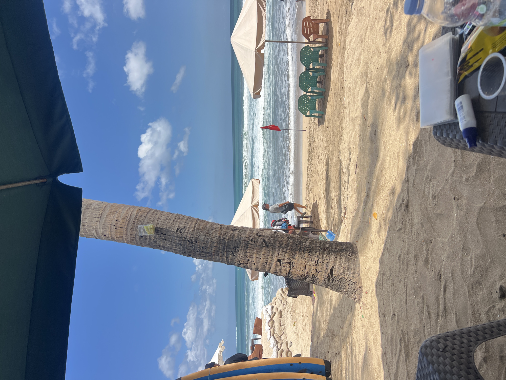 The image depicts a beach scene in Kuta, Indonesia, on a clear, sunny day. In the foreground, a large palm tree trunk dominates the left side, with some debris on its bark. Part of a dark-colored outdoor chair and a table are visible in the lower left corner. The table holds a clear plastic bottle of water, a white tube of sunscreen, a clear plastic cup, and a yellow package. Sand covers the ground, with some shadows cast by unseen objects.
In the middle ground, the sandy beach stretches towards the ocean. Several beach chairs, some green and some orange, are placed under a tan umbrella. A small red flag stands near the water's edge. The ocean features moderate waves with whitecaps, indicating active surf. Two individuals are walking on the beach near the water, and another person is seen further back near some sandbags and what appear to be more beach seating. Surfboards are leaning against a wall or structure on the far left. The sky is a vibrant blue with scattered white clouds. The overall atmosphere is one of a relaxed beach day.